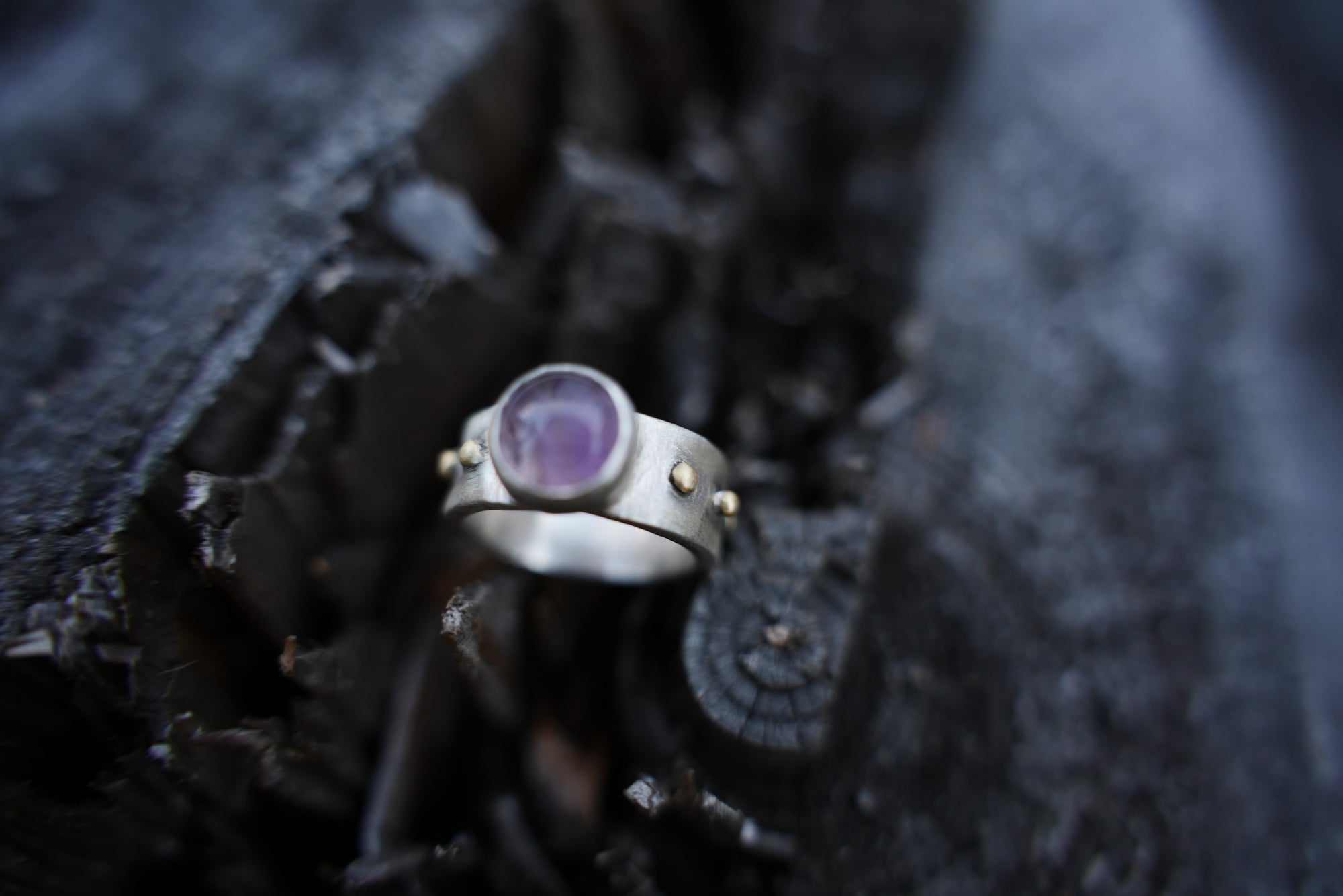 A sterling silver and 18K gold ring with a purple amethyst gemstone against a dark, blurred background.