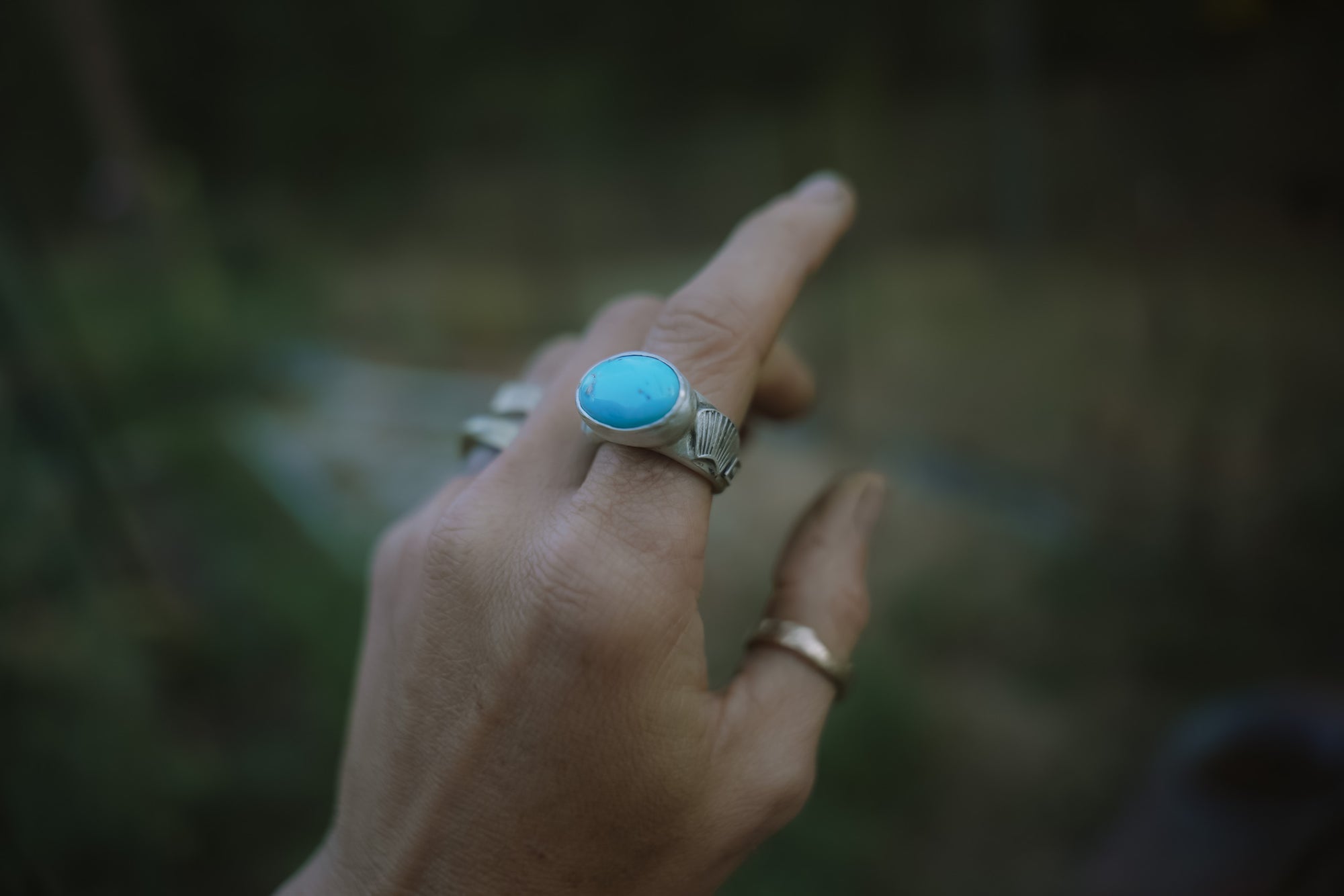 Hand wearing a sterling silver ring with a hand-stamped band and a turquoise stone against a blurred natural background