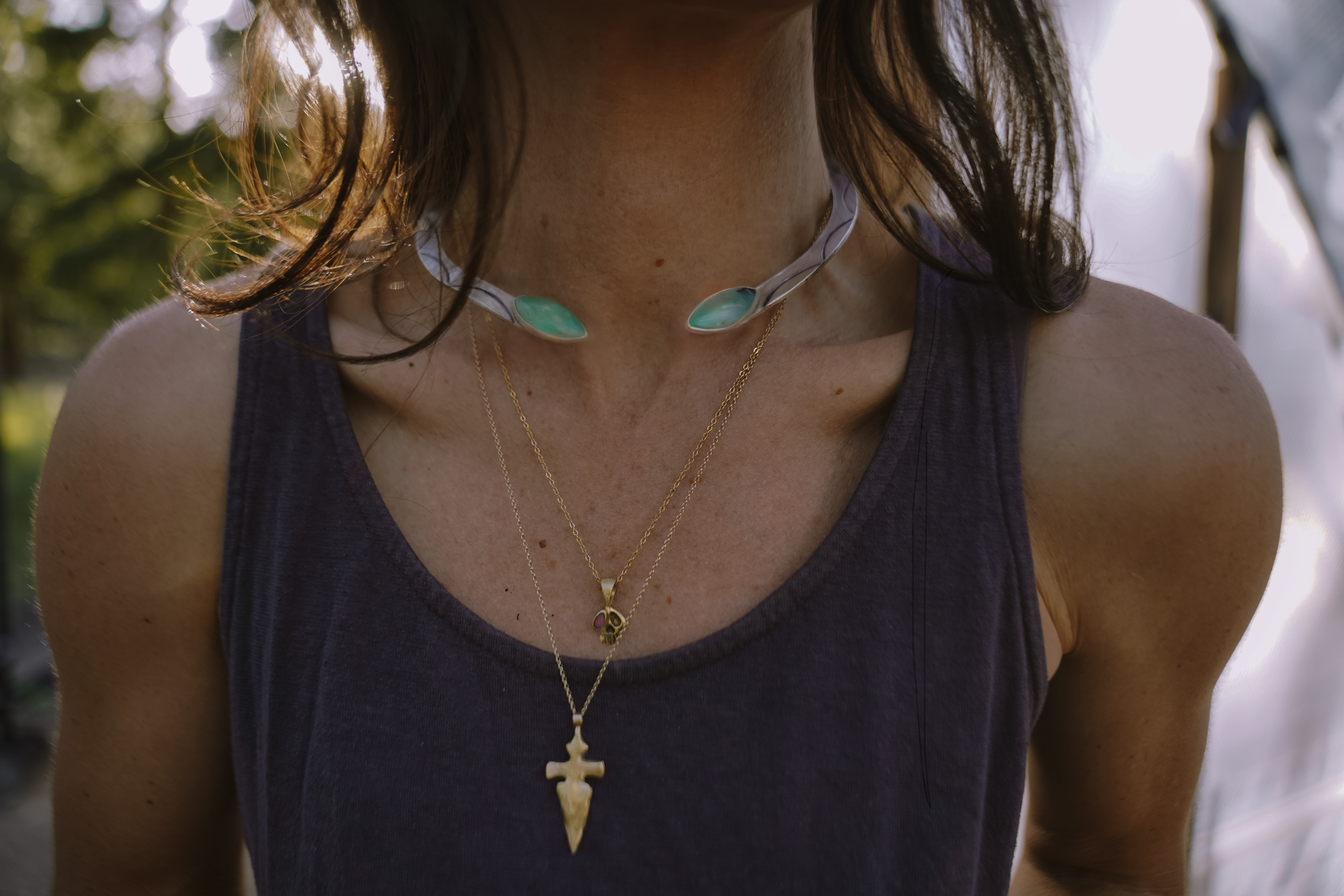Person wearing multiple gold necklaces and a silver choker against a blurred background. Necklace charms include a gold dagger and a gold skull with a pink tourmaline accent.