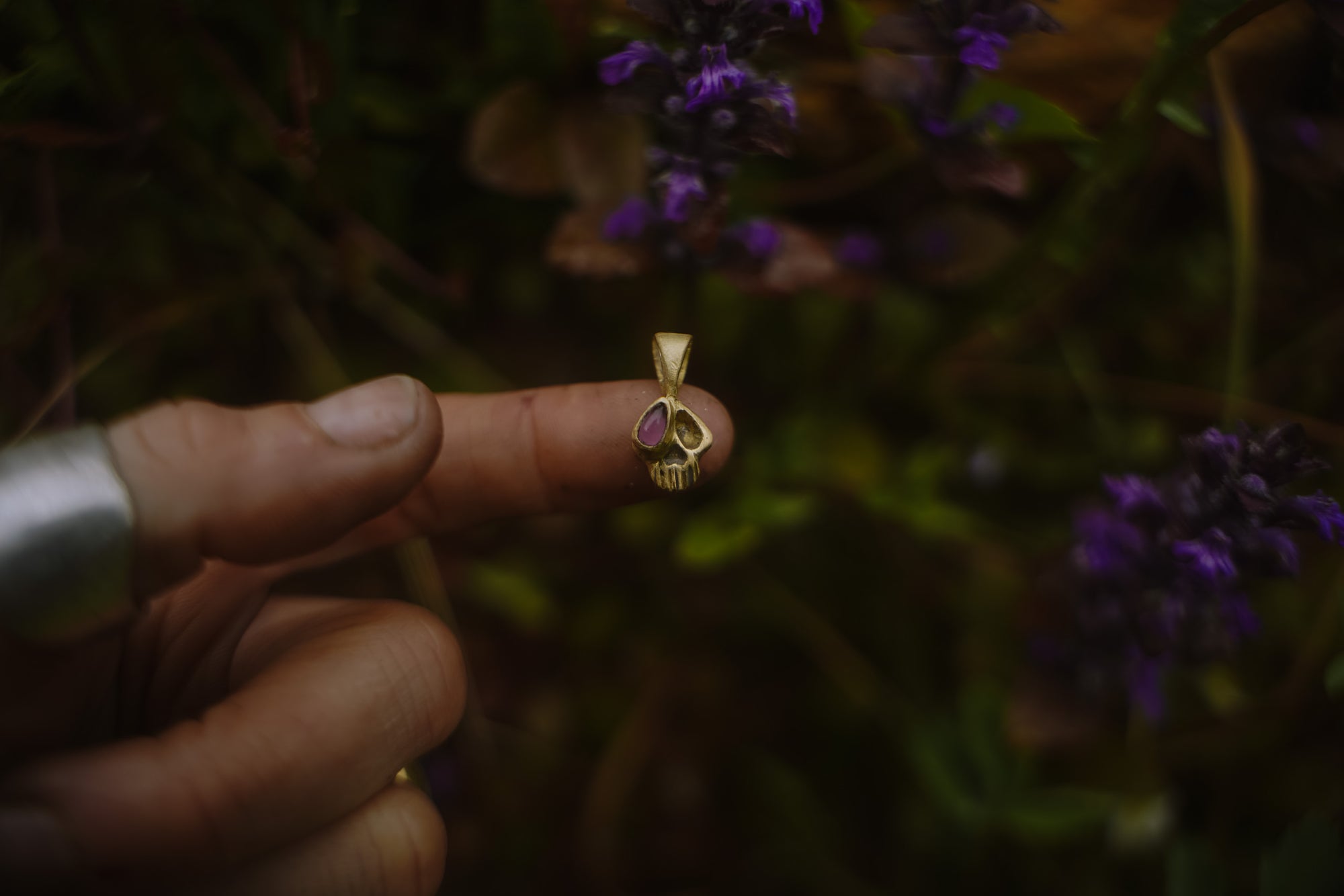 18K gold Momento Mori skull pendant with pink tourmaline accent held between fingers against a blurred natural background