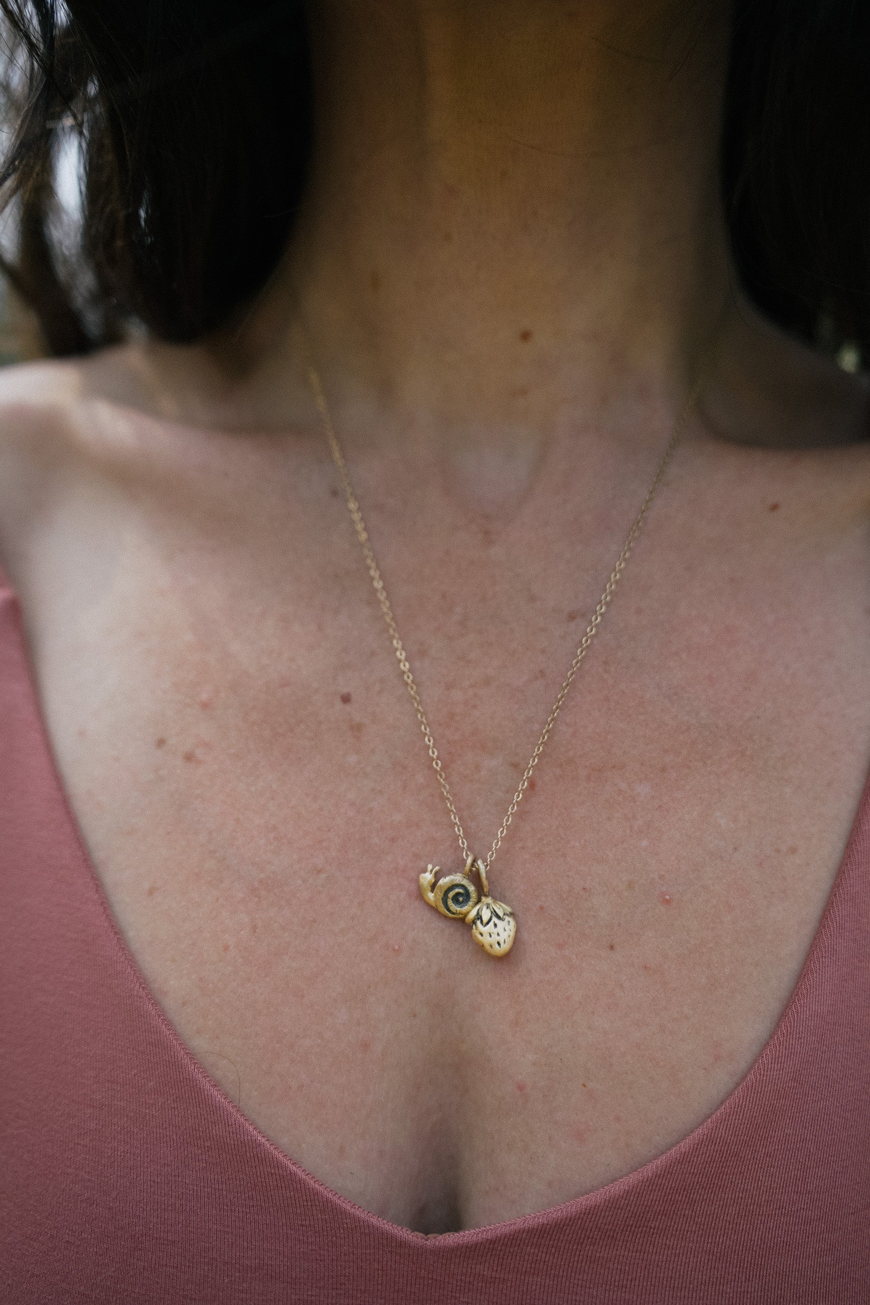 Gold necklace with a snail charm and a strawberry charm worn by a person in a pink top.