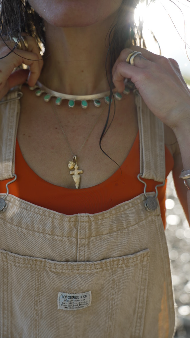 A woman wearing a necklace with a gold strawberry charm and dagger pendant. She is wearing tan overalls and an orange top.