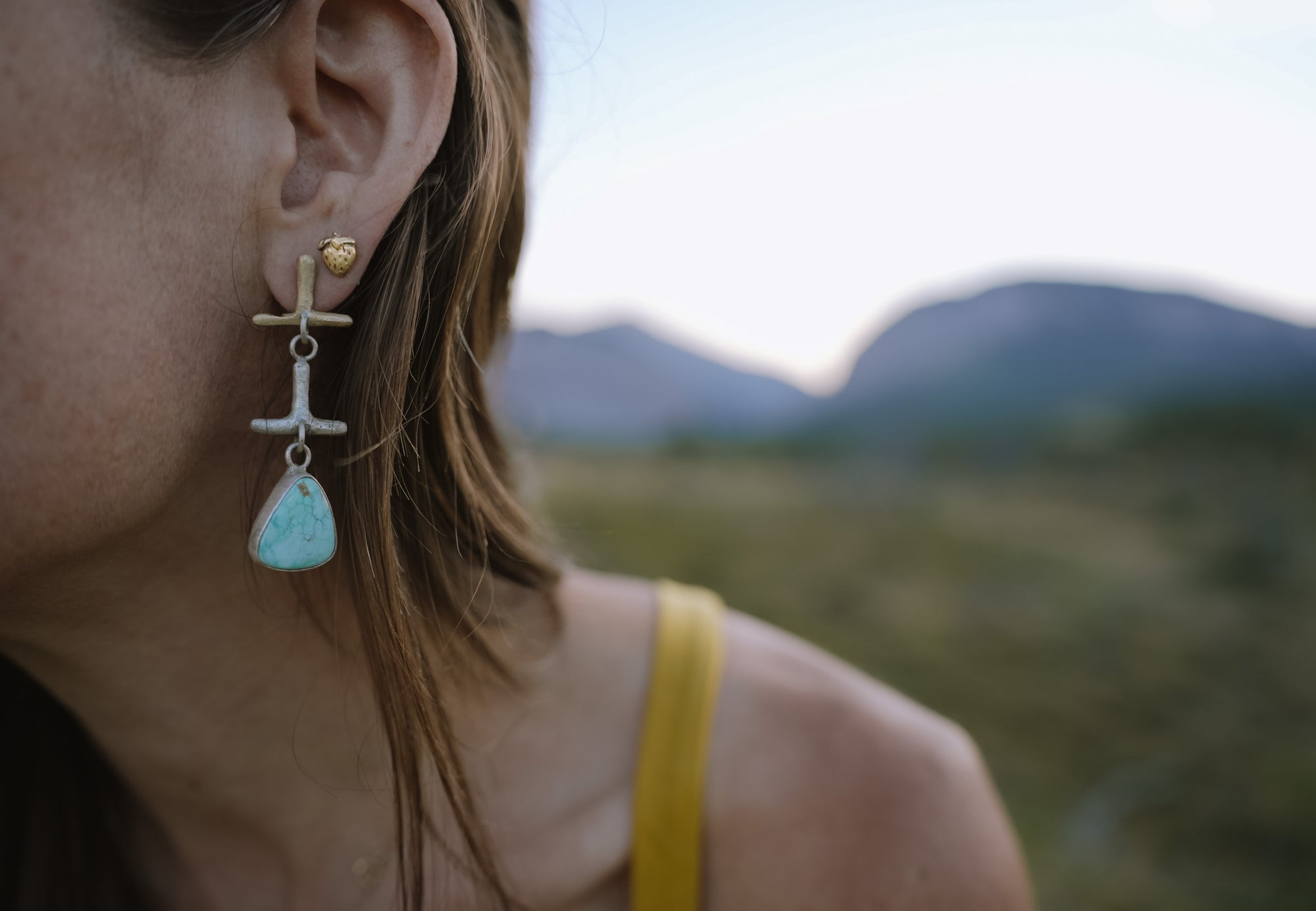 Close-up of a person wearing a gold strawberry-shaped post earring and a dangling silver and gold earring with turquoise, with a blurred natural background .