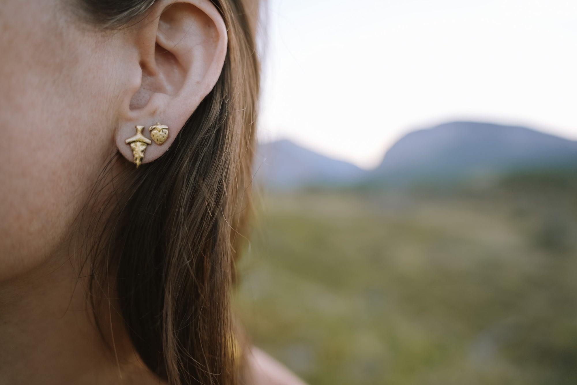 Close-up of a person wearing a gold strawberry-shaped earrings and a gold dagger-shaped earring with a blurred natural background