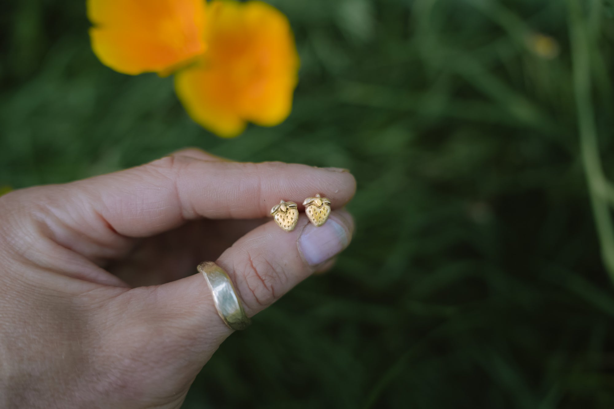 Hand holding gold strawberry-shaped earrings with blurred flowers in the background.