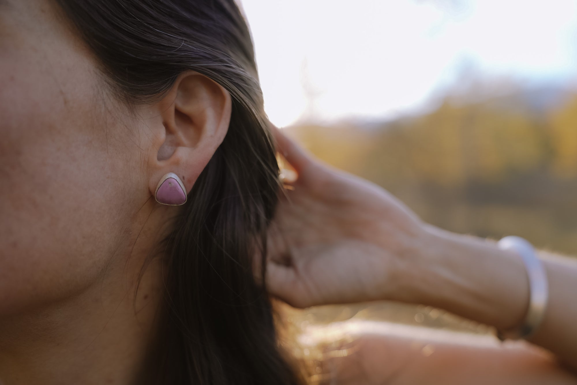 A woman wearing sterling silver and pink phosphosiderite post-style , triangular-shaped earrings against a blurred natural background.