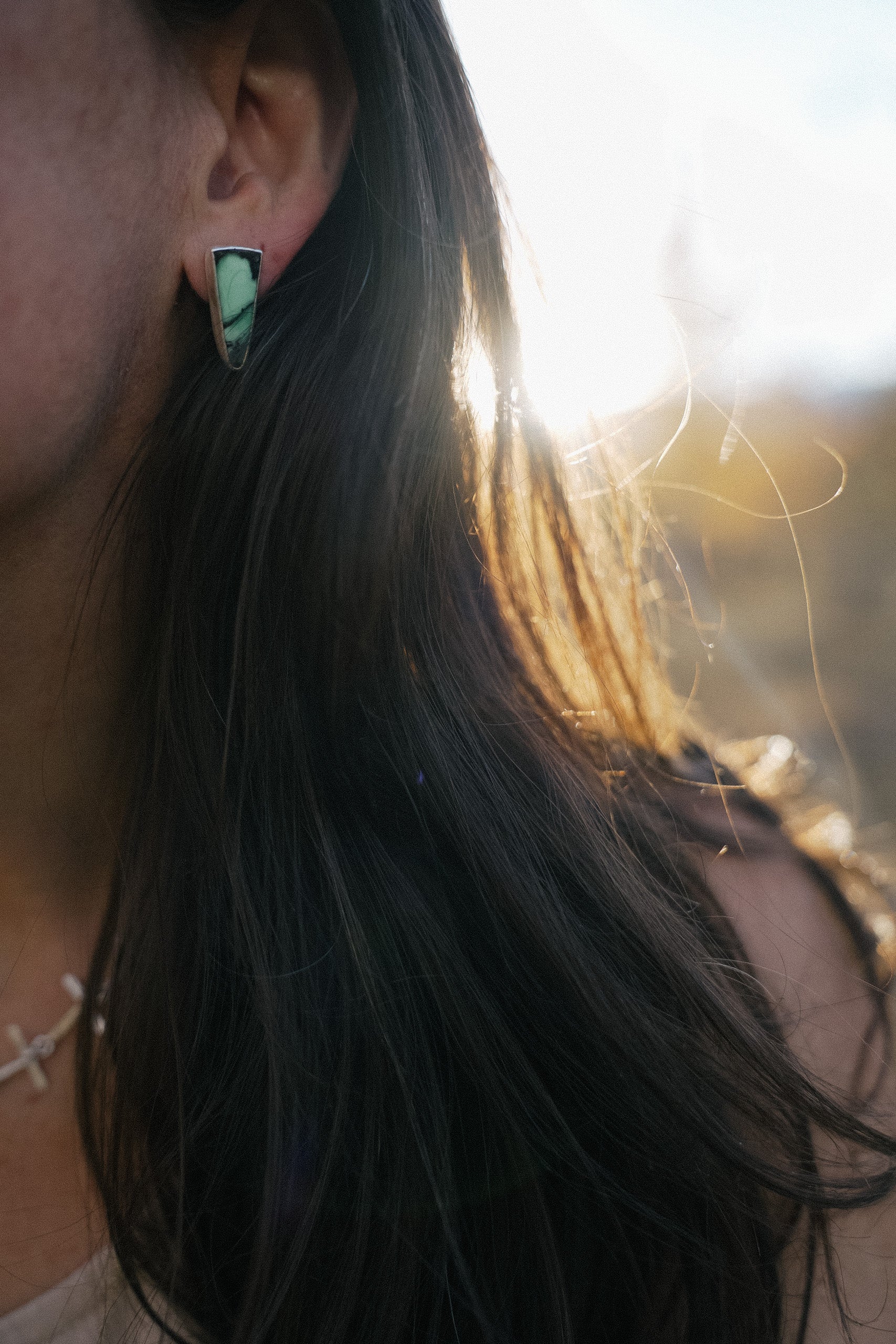 Close-up of a woman's ear with a silver and green gemstone spike-shaped earring against a blurred natural background.