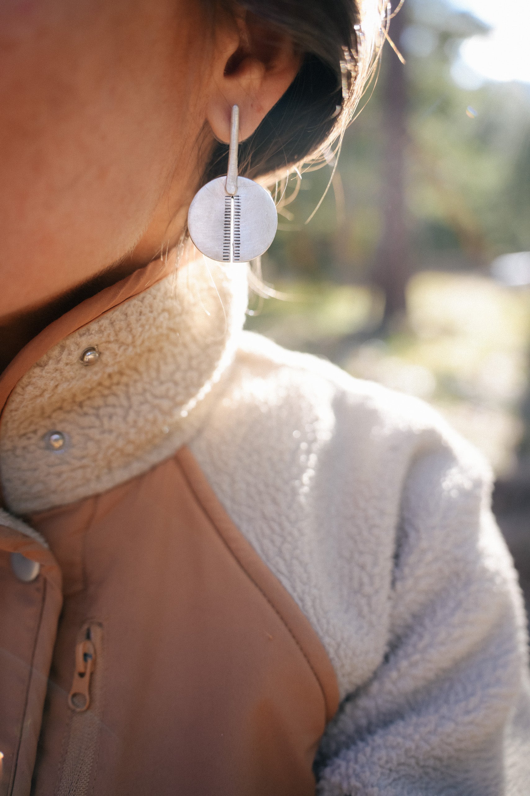 Close-up of a woman with ldark hair wearing a sterling silver disc-shaped, hand-stamped earring. The disk has a cut running from the bottom almost to the top with short, horizontal lines radiating from each side of the cut. There is a narrow, silver section connecting the disk to her ear lobe. The natural sun-lit background is blurred.