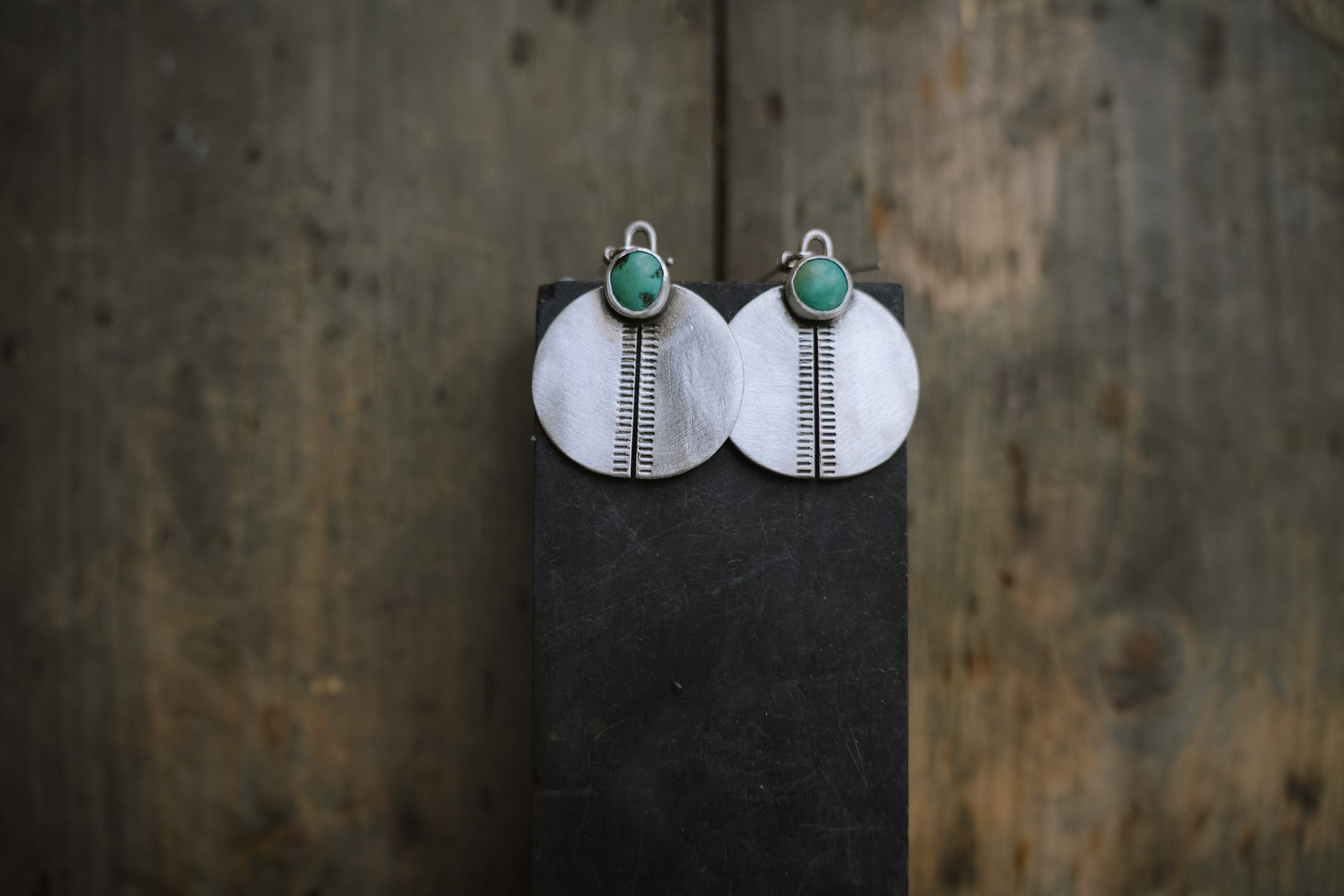 Pair of sterling silver disc-shaped and hand-stamped earrings with small green turquoise gemstones at the top. Set on a dark surface.