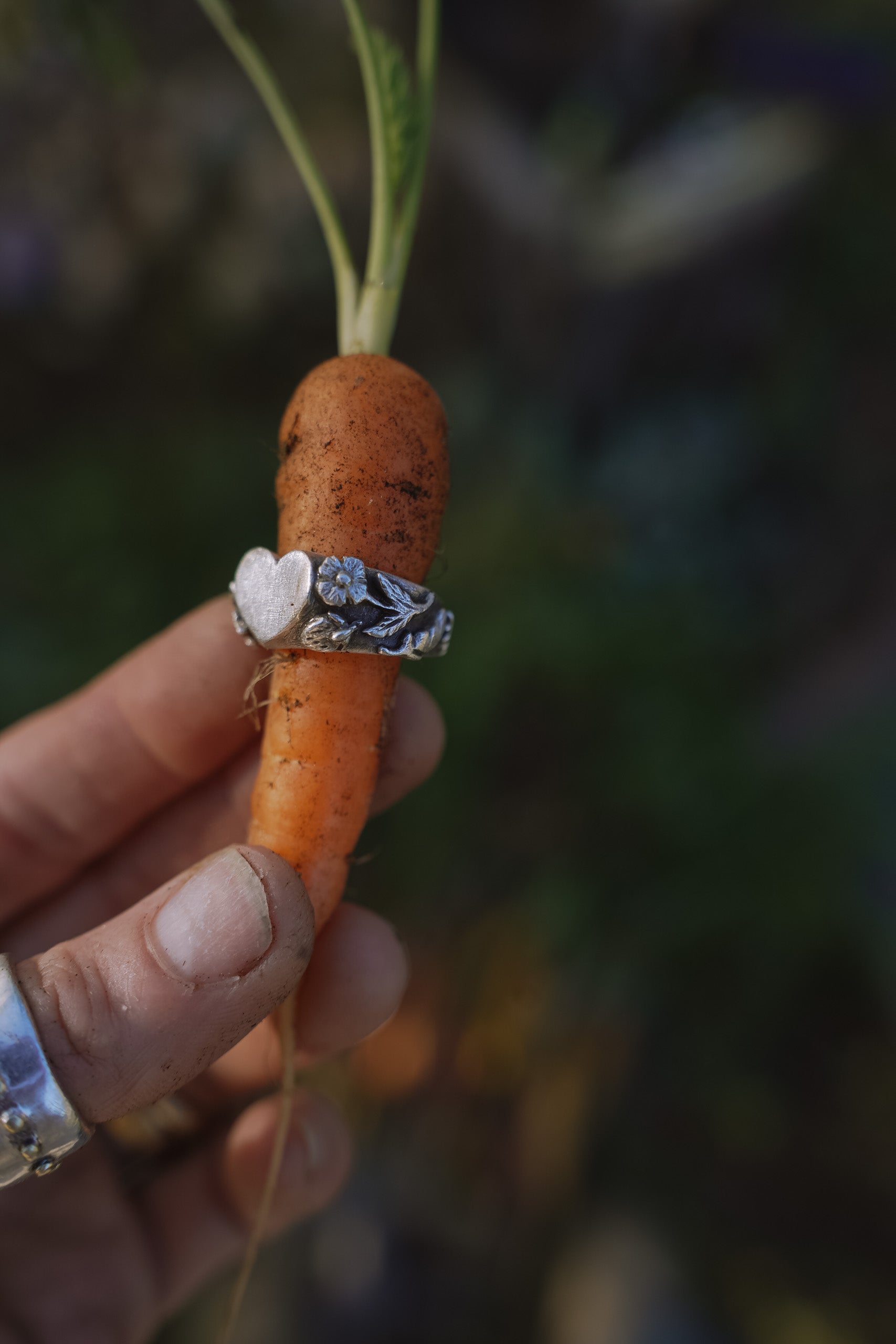 Sterling silver signet ring with a heart shape at the top and carved floral motifs on the band. Shown on a carrot held by a woman's hand against a natural background.