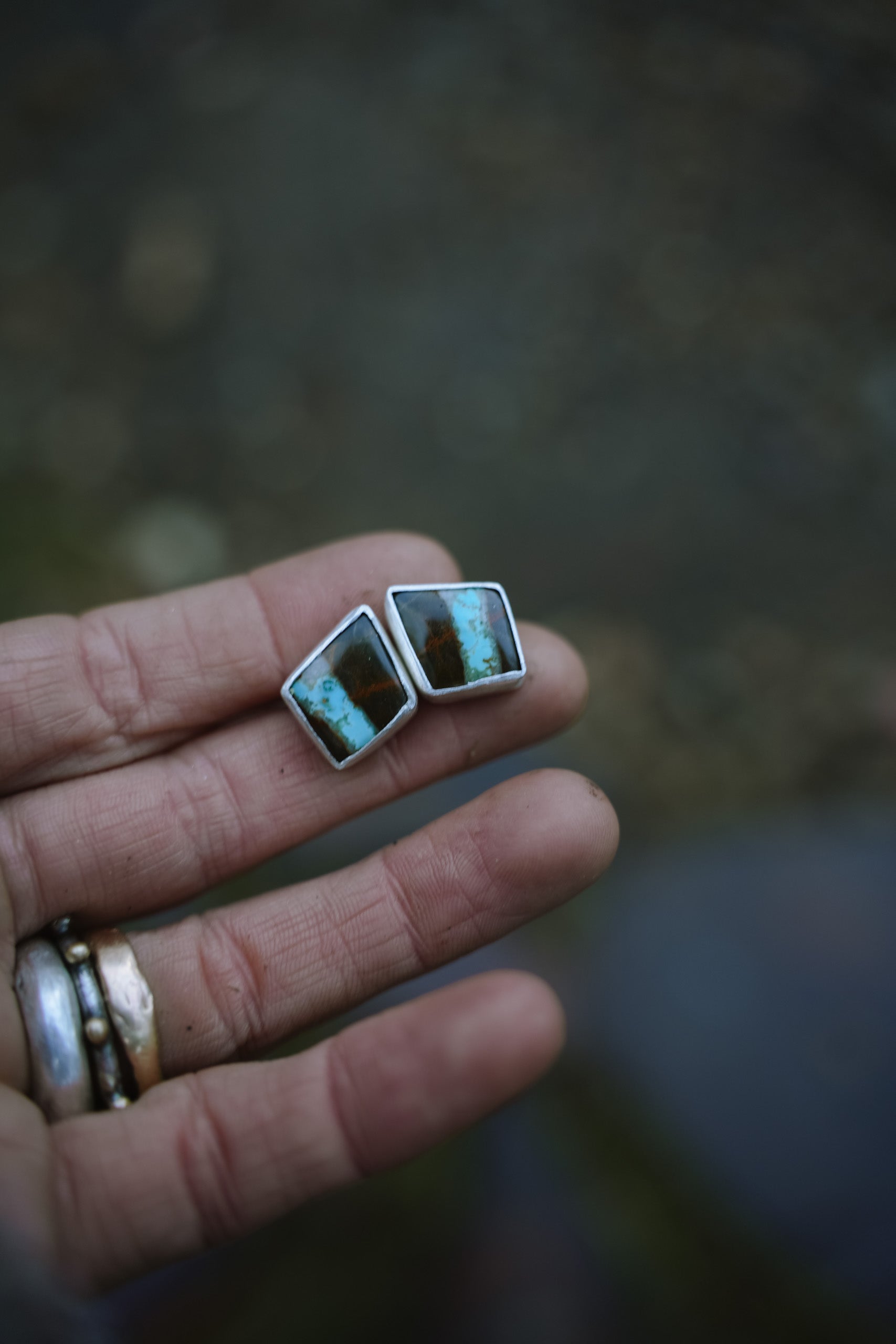 Triangular-shaped earrings with two-toned blue and black chrysolla gemstones set in sterling silver and held in a woman's hand against a blurred natural background