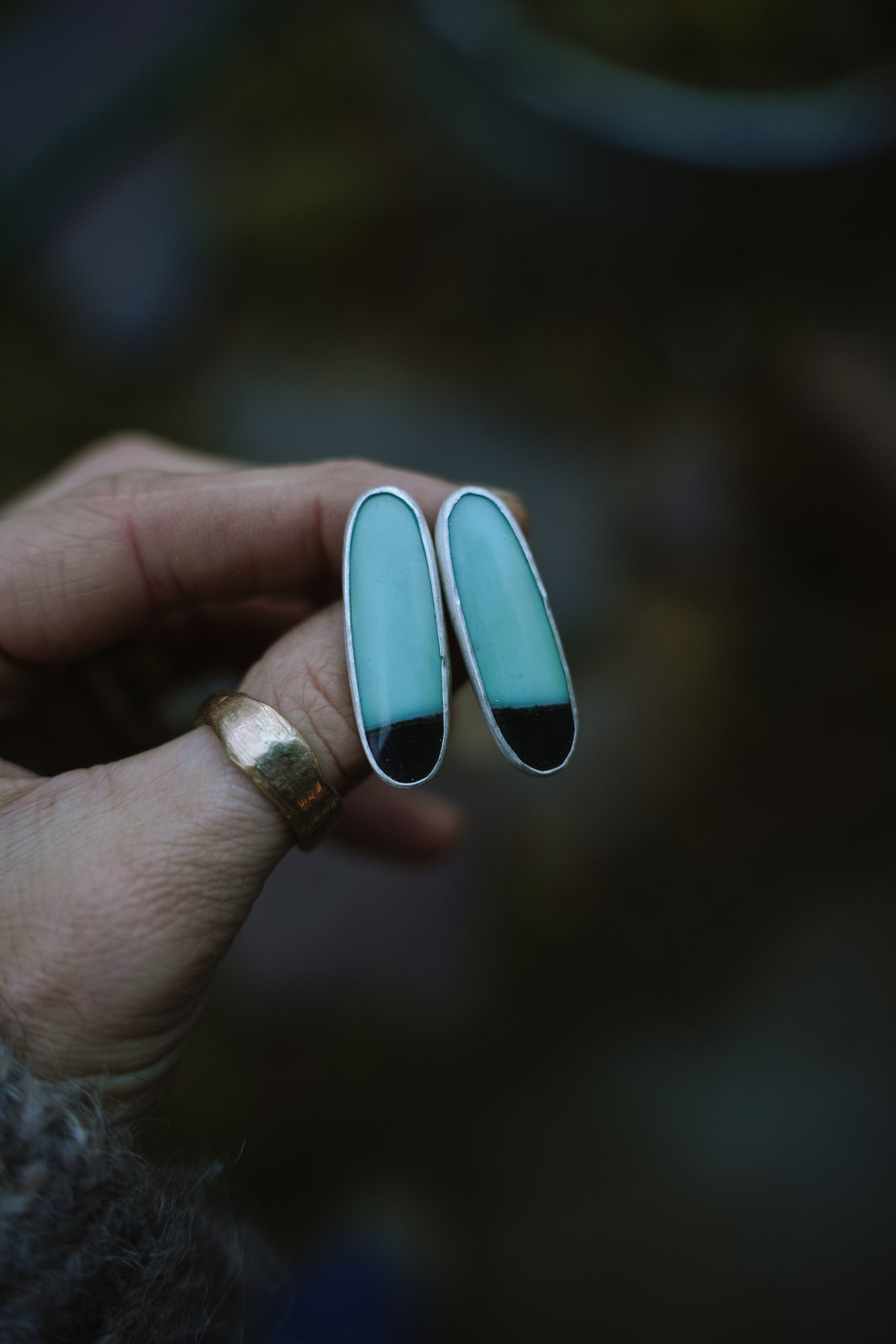 Long, slim, oval-shaped earrings with two-toned blue and black chrysolla gemstones set in sterling silver and held in a woman's hand against a blurred natural background.