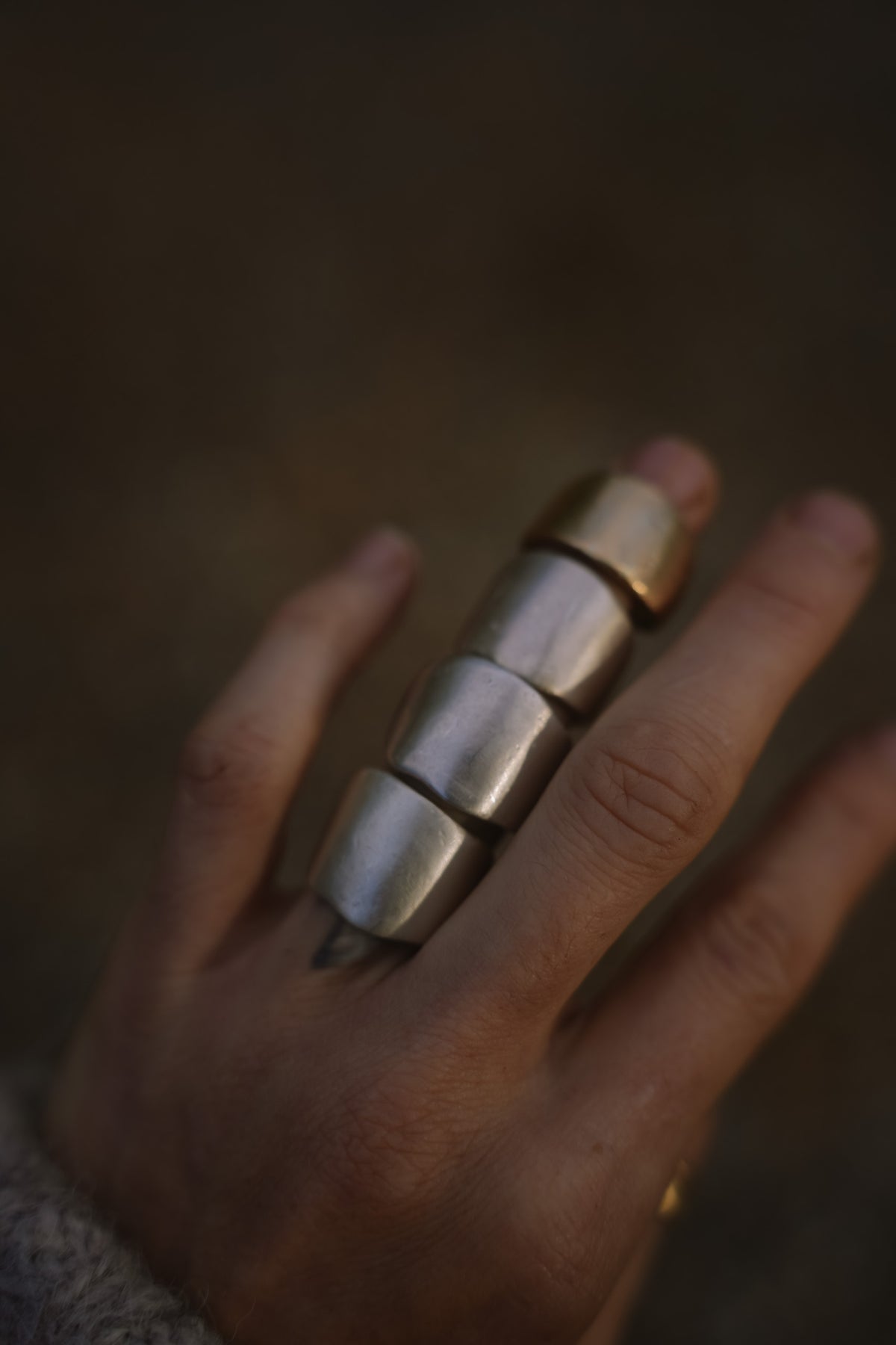 Close-up of a woman's hand with four wide, smooth band rings stacked along one finger, three in a soft matte silver tone and the topmost in warm gold, against a dark, muted brown background. A gray knit sleeve is just visible at the lower left.