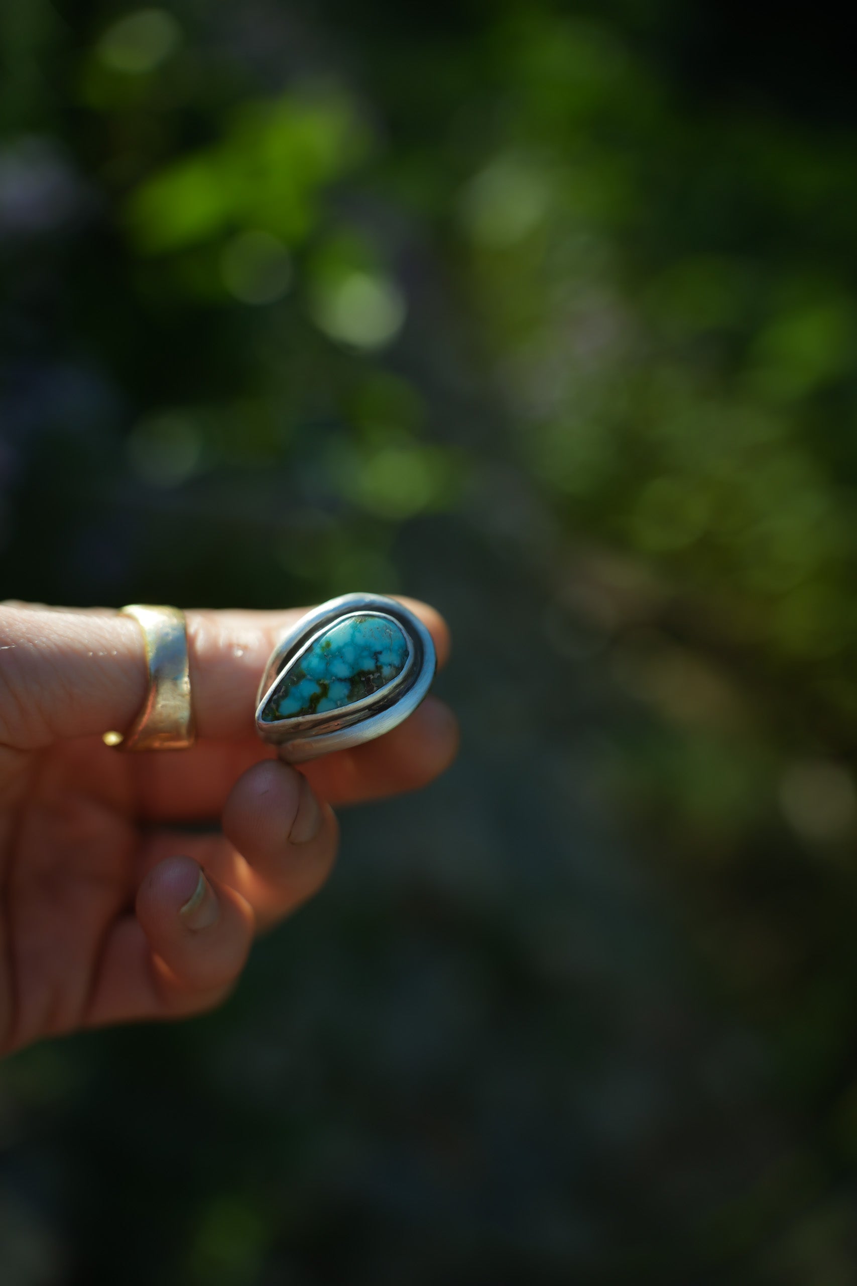 A woman's hand holding a large and bold sterling silver ring with a teardrop-shaped turquoise gemstone, set against a blurred green outdoor background.