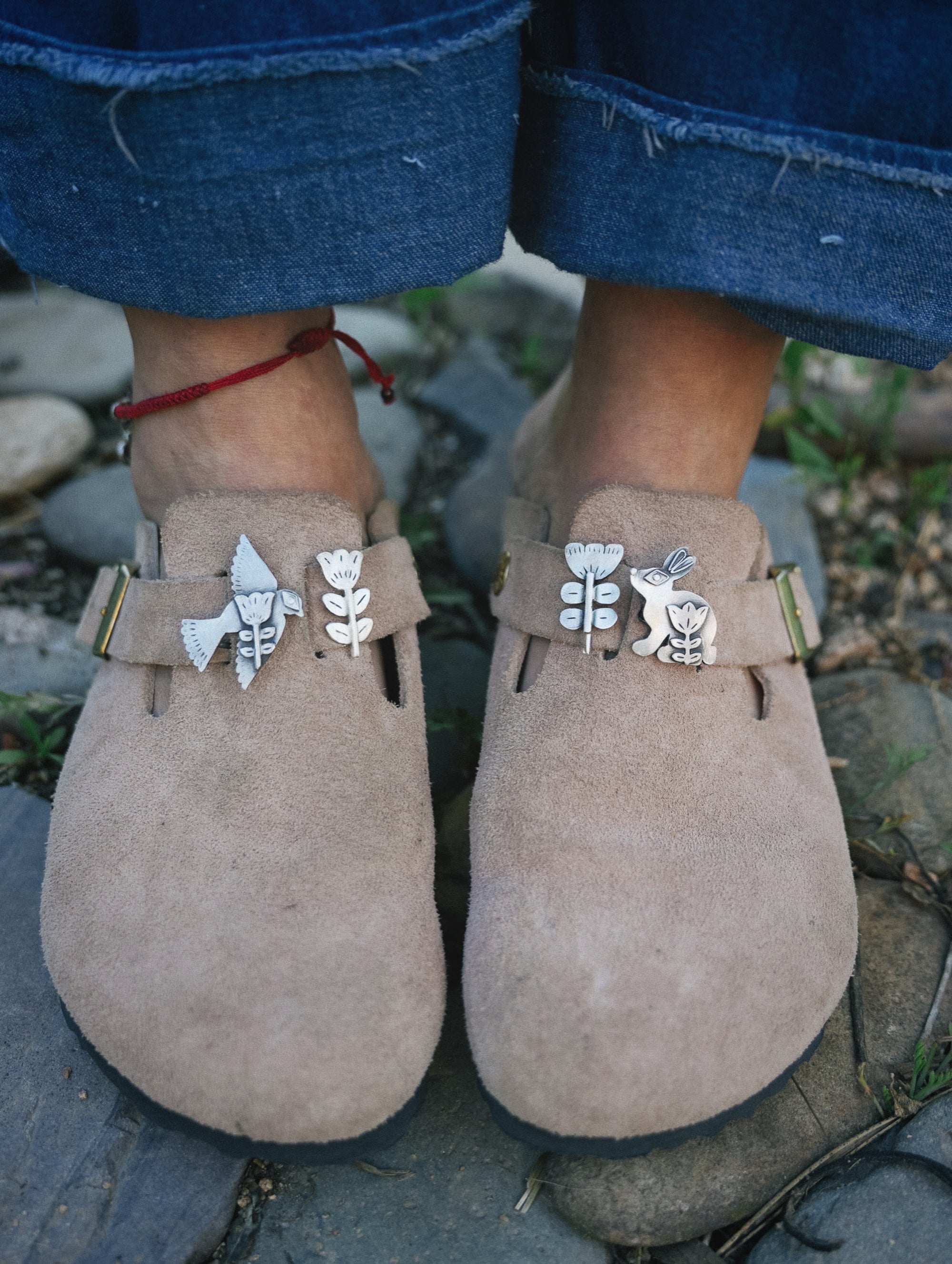A woman wearing light brown suede Birkenstock shoes with decorative, folksy silver buckles attached. She is standing on a stone surface.