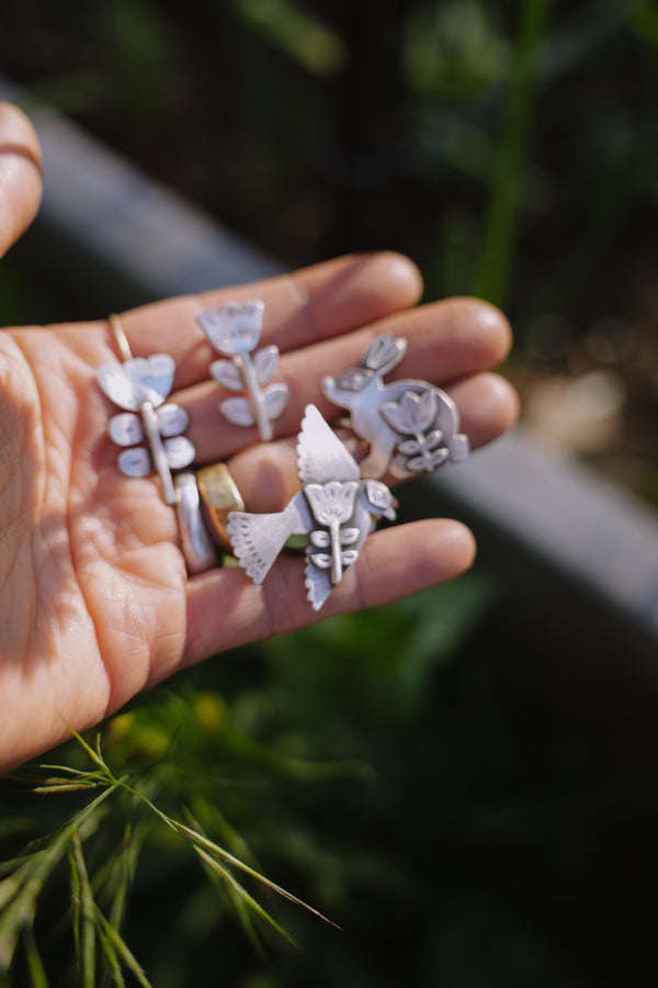 A woman's hand holding four folk-style sterling silver floral Birkenstock shoe clips against a natural background.