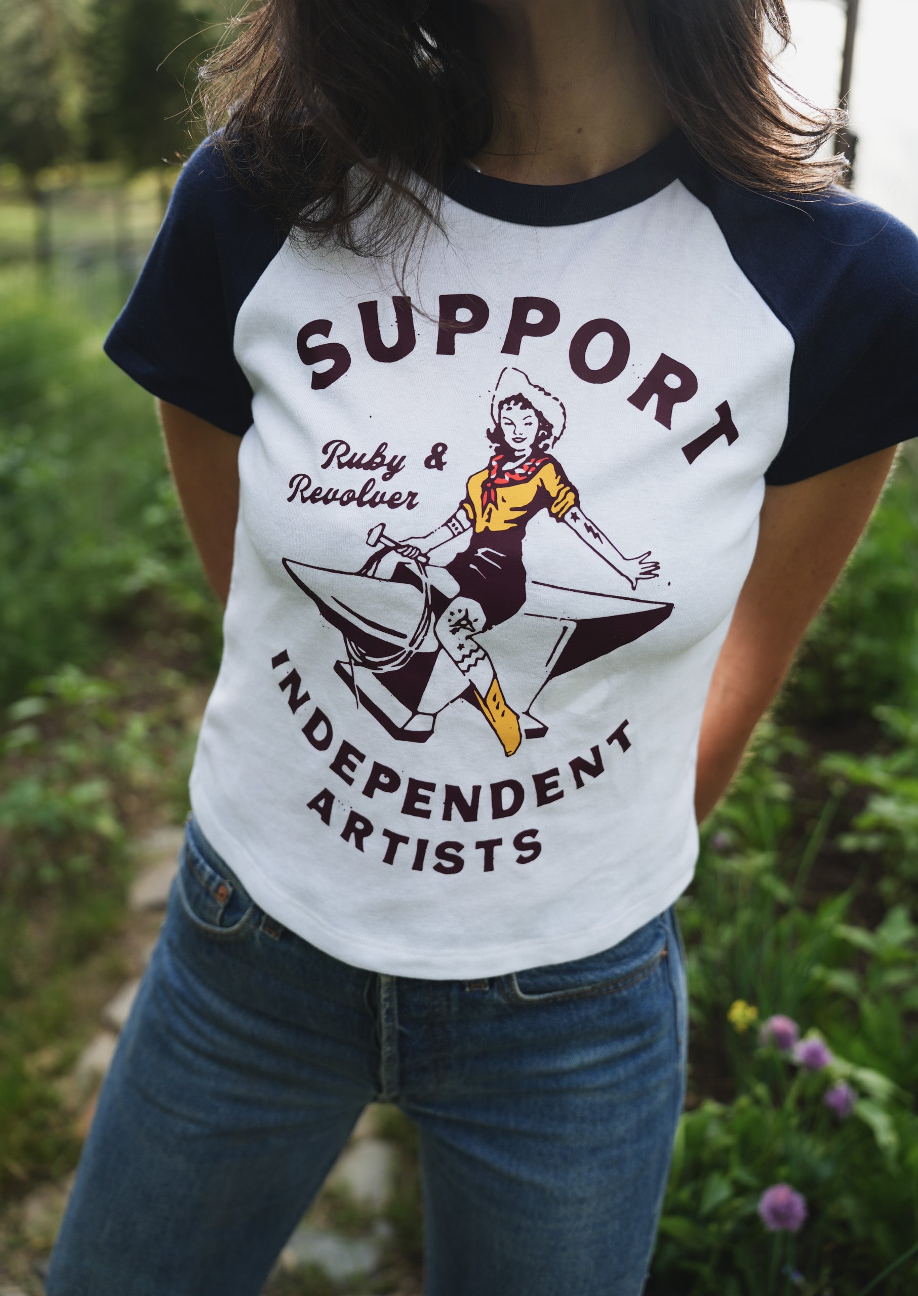 A woman with long, dark hair wearing a white and navy raglan t-shirt with a vintage-style graphic of a cowgirl riding an anvil, surrounded by the text 'Ruby & Revolver  SUPPORT INDEPENDENT ARTISTS'. The background is a blurred outdoor garden setting.