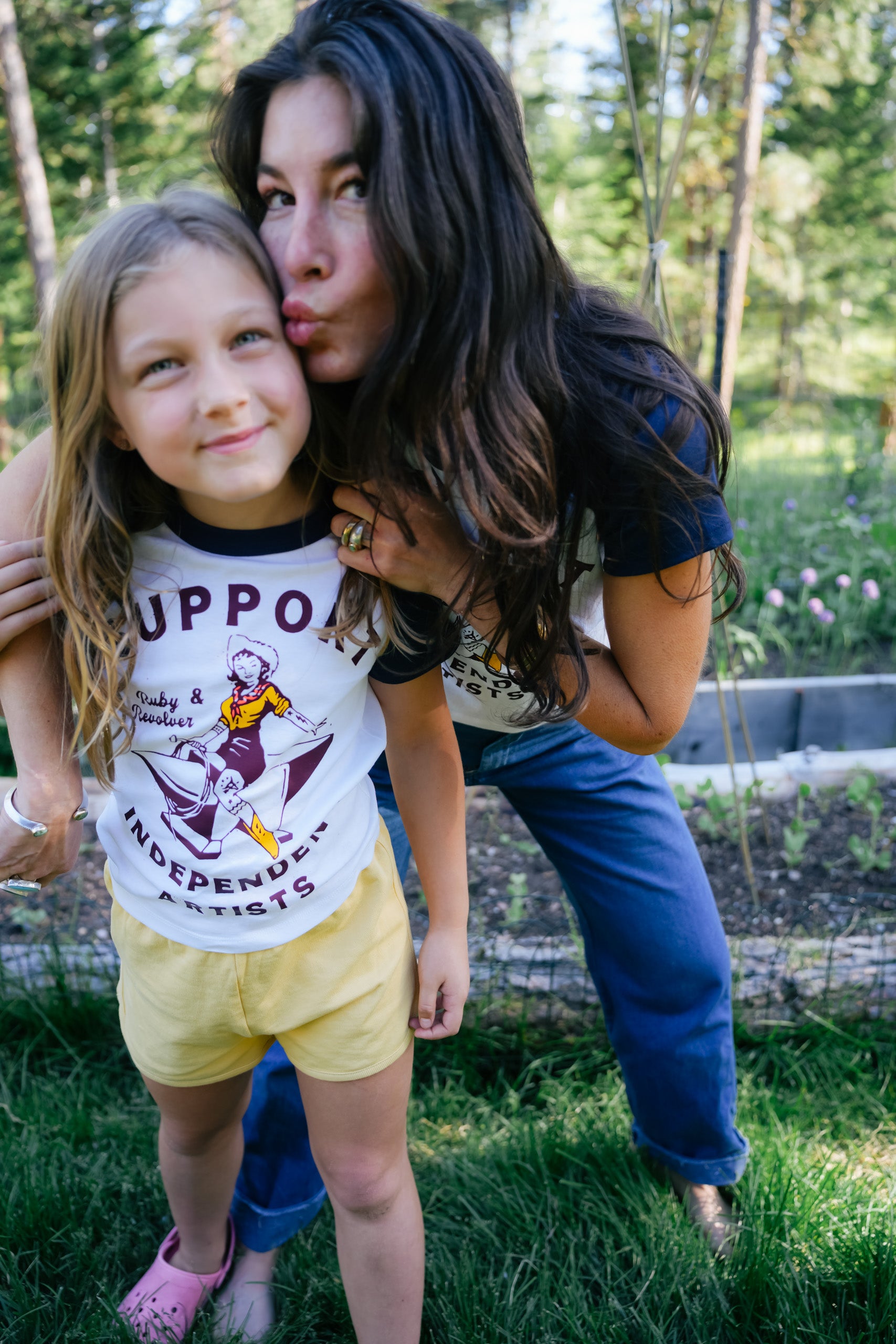 A woman with long, dark hair kissing the cheek of a young girl. She is wearing a white and navy raglan t-shirt with a vintage-style graphic of a cowgirl riding an anvil, surrounded by the text 'Ruby & Revolver  SUPPORT INDEPENDENT ARTISTS'. The background is a blurred outdoor garden setting.