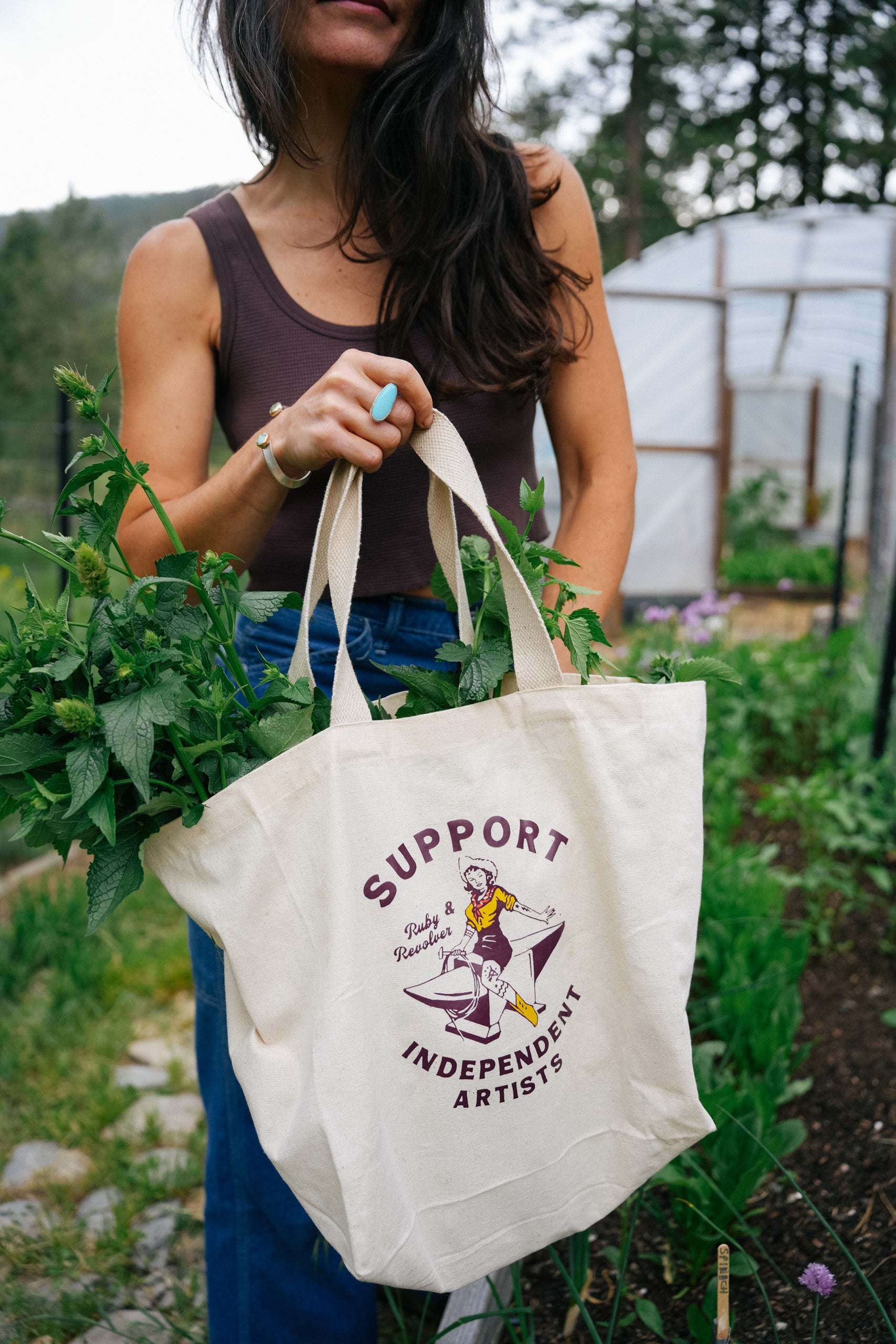A woman with dark hair holding a white cotton tote back with a vintage-style logo. The logo shows a cowgirl riding an anvil and say “Support Independent Artists.” She is standing in a vegetable garden with raised beds in front and a hoop house in the background.