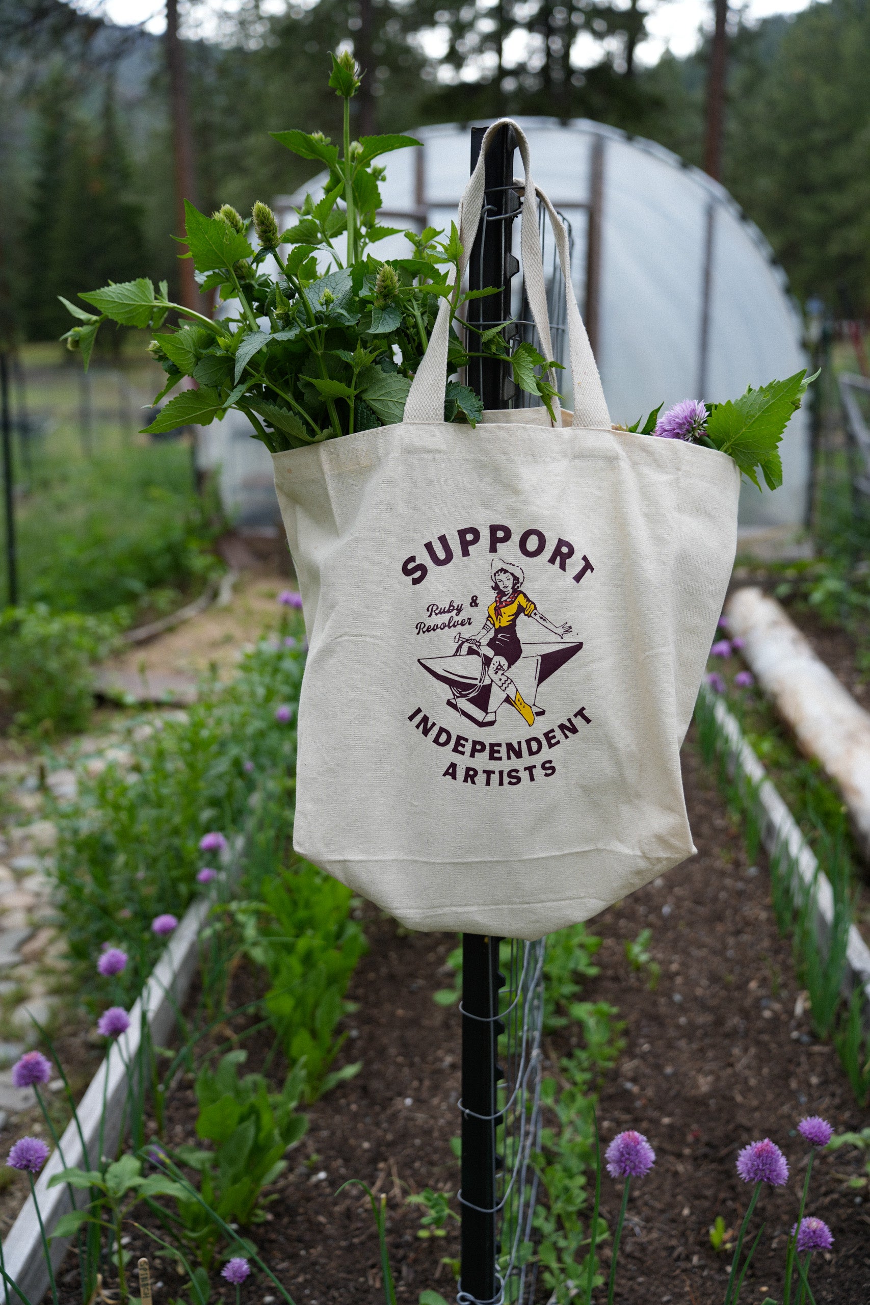 A white cotton tote back with a vintage-style logo. The logo shows a cowgirl riding an anvil and say “Support Independent Artists.” The tote is handing from the edge of a post in a vegetable garden raised bed. There is a hoop house in the background.