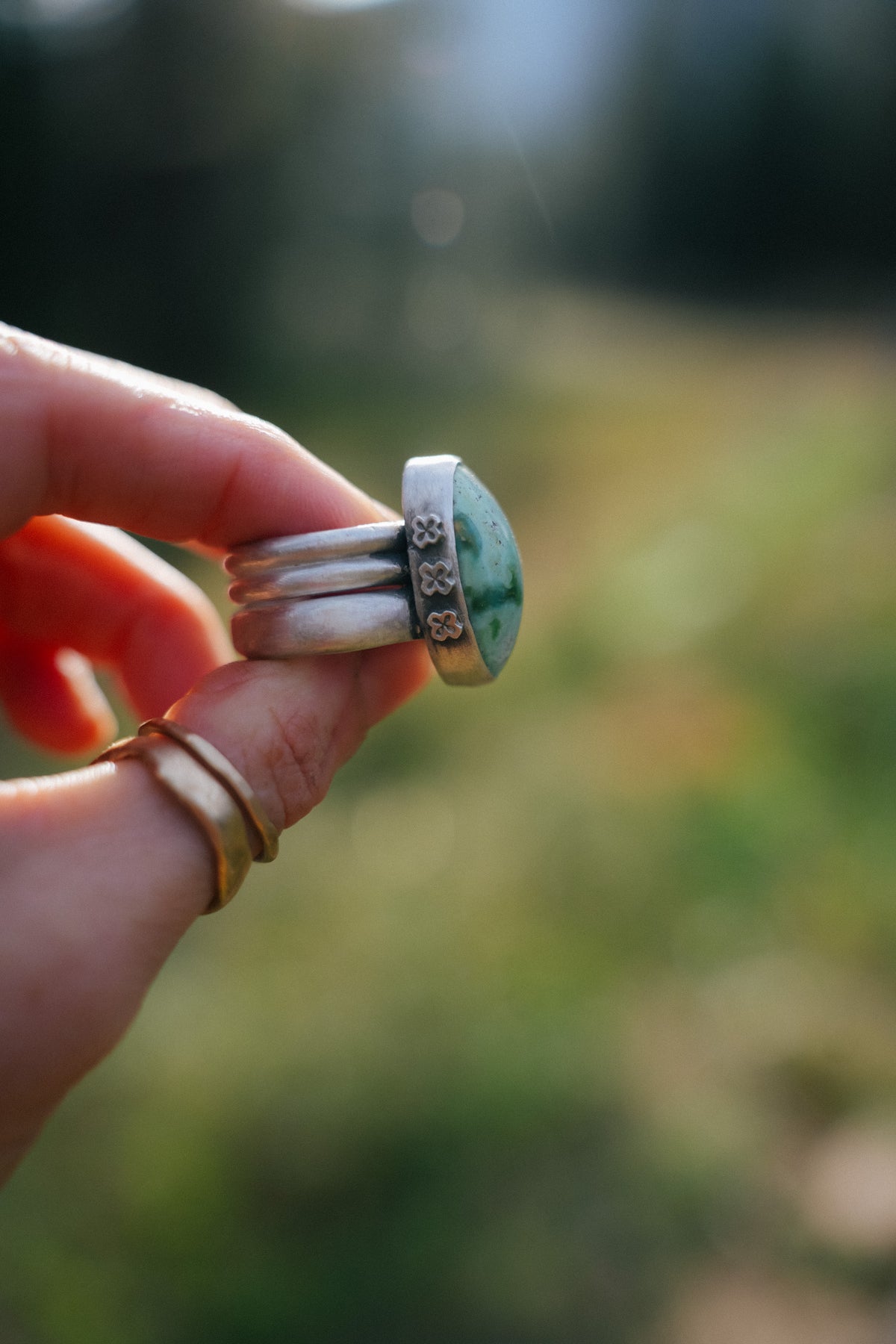 Side view of the Cruces ring held between two fingers, showing a Royston turquoise stone in a bezel stamped with a row of small four-petal flowers (stars or crosses), above an extra wide sterling silver band made of three stacked ridges. Two thin gold bands sit on the finger below, against a blurred green and golden background.