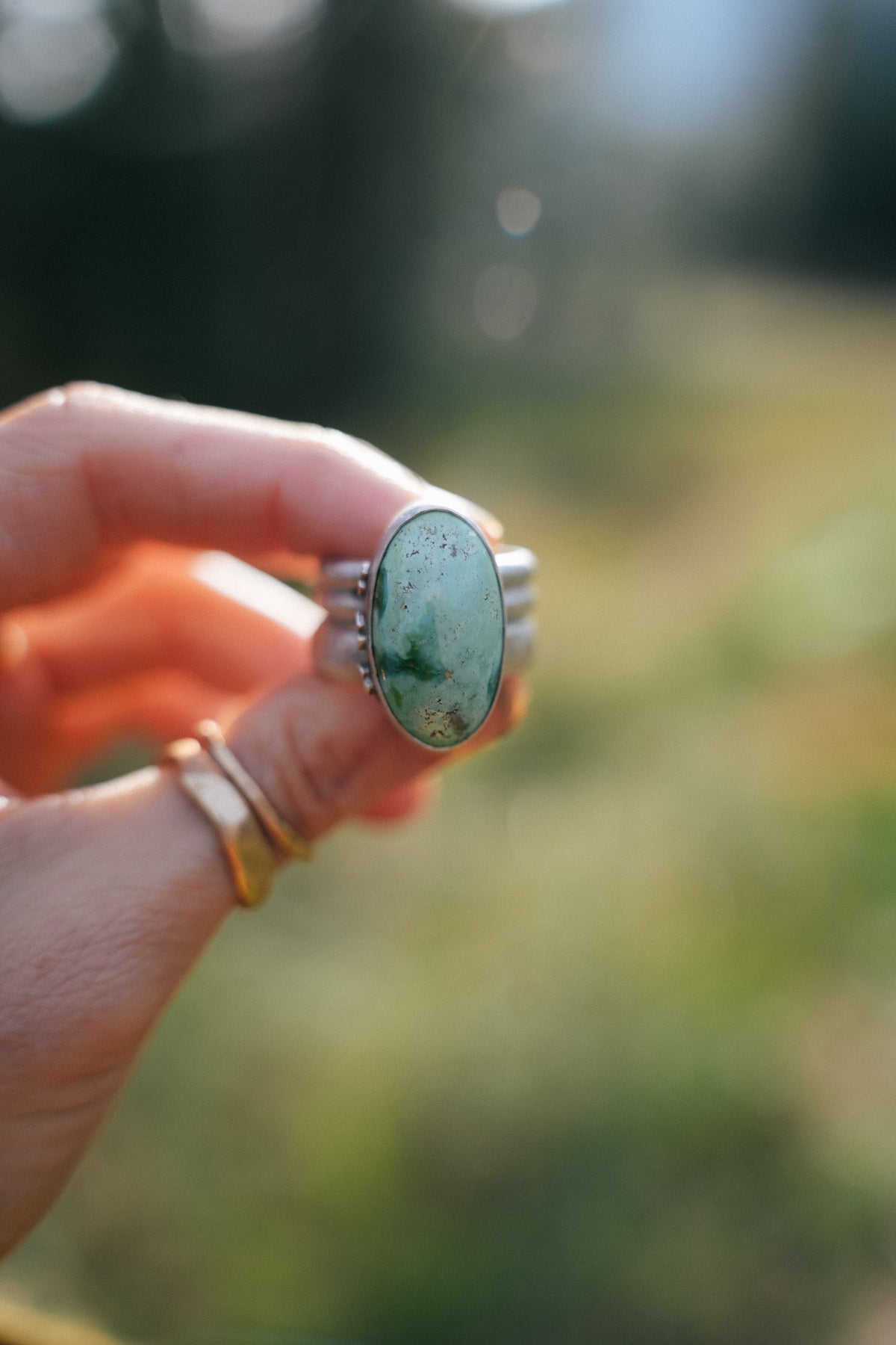 Front view of the Cruces ring held between two fingers, with a large oval Royston turquoise gemstone in a smooth silver bezel. The soft green stone has darker green swirls and fine brown flecks, and small silver beads decorate the sides of the extra wide ridged band. Two thin gold bands are stacked on the finger below, against a blurred green and golden background.
