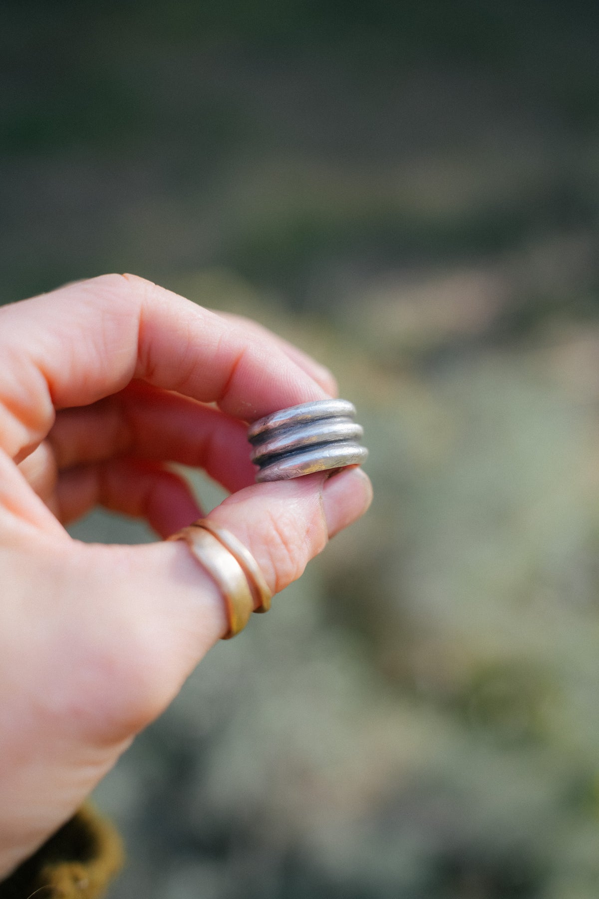 Close-up of the extra wide band of the Cruces ring, three rounded, slightly oxidized sterling silver ridges stacked together, held between a thumb and finger with two thin gold bands below, against a blurred green background.