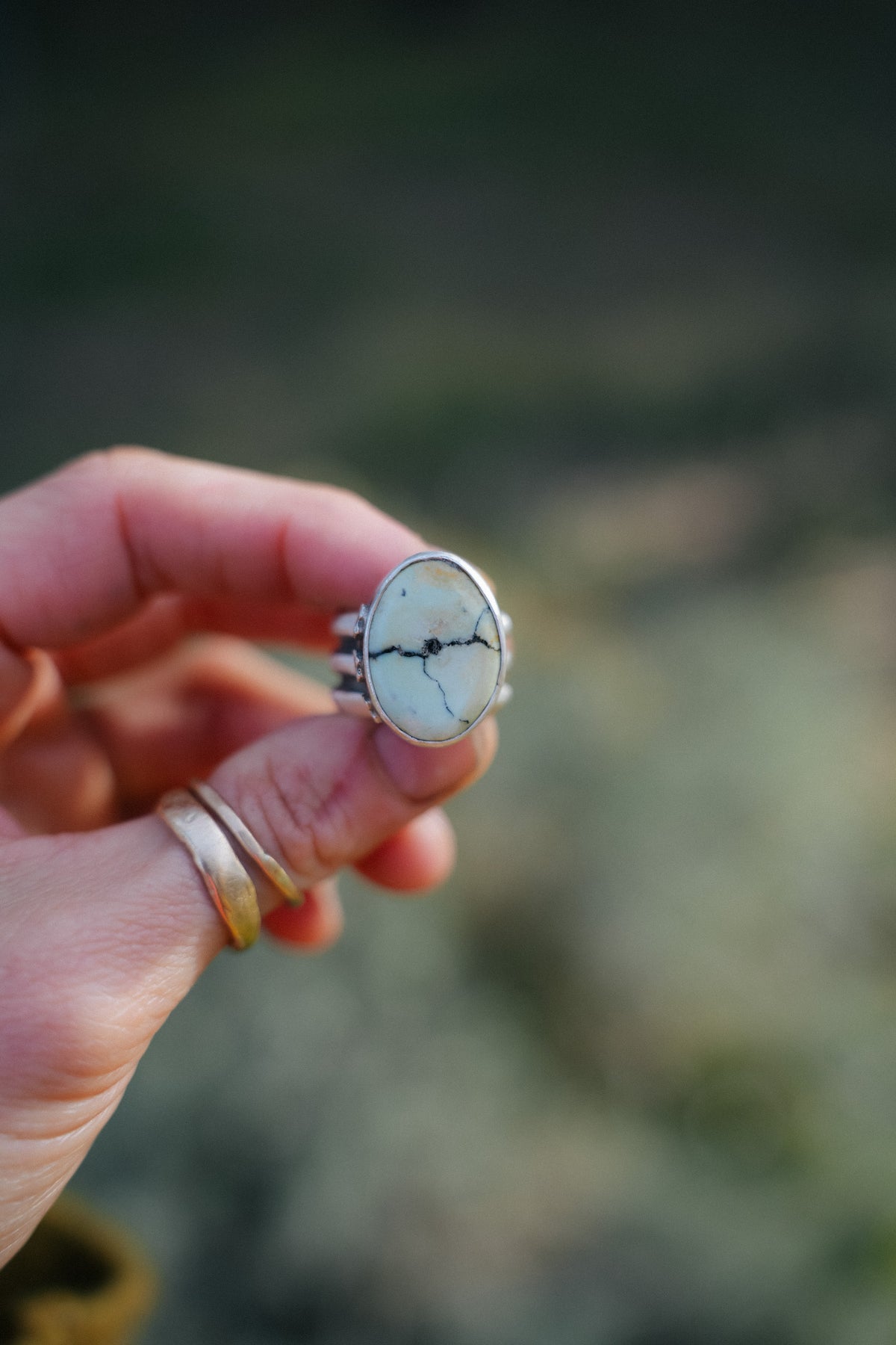 Cruces ring, a sterling silver ring with an oval pale green-white Aloe variscite stone crossed by thin black veins, held up between two fingers with two thin gold bands stacked on the finger below, against a soft blurred green background.