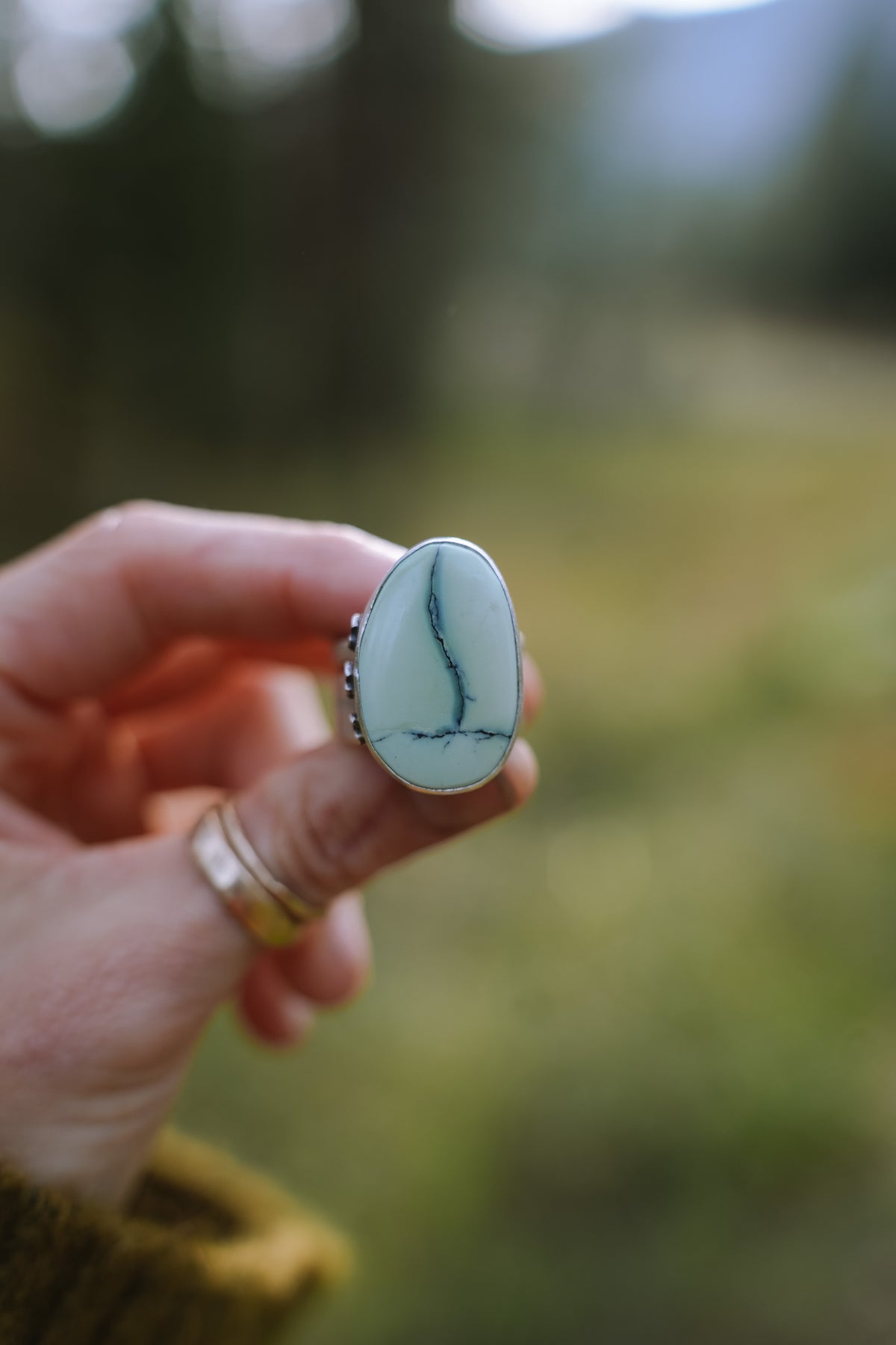 Front view of the Cruces ring held between two fingers, with a large pale blue-green Aloe variscite stone in a smooth silver bezel. A dark vein runs down the stone and branches into a thin horizontal line near the bottom. Two thin gold bands are stacked on the finger below, against a soft green blurred background.