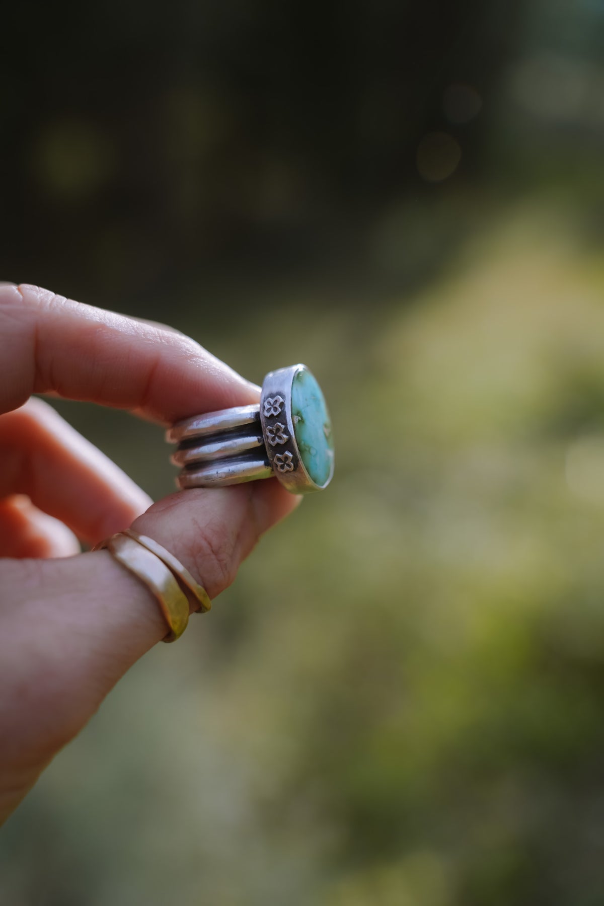 The Cruces ring seen from the side, held between two fingers, showing a Sonoran Gold turquoise stone in a bezel stamped with a row of small four-petal flowers or crosses/stars, above an extra wide sterling silver band made of three stacked ridges. Two thin gold bands sit on the finger below, against a blurred green and golden background.