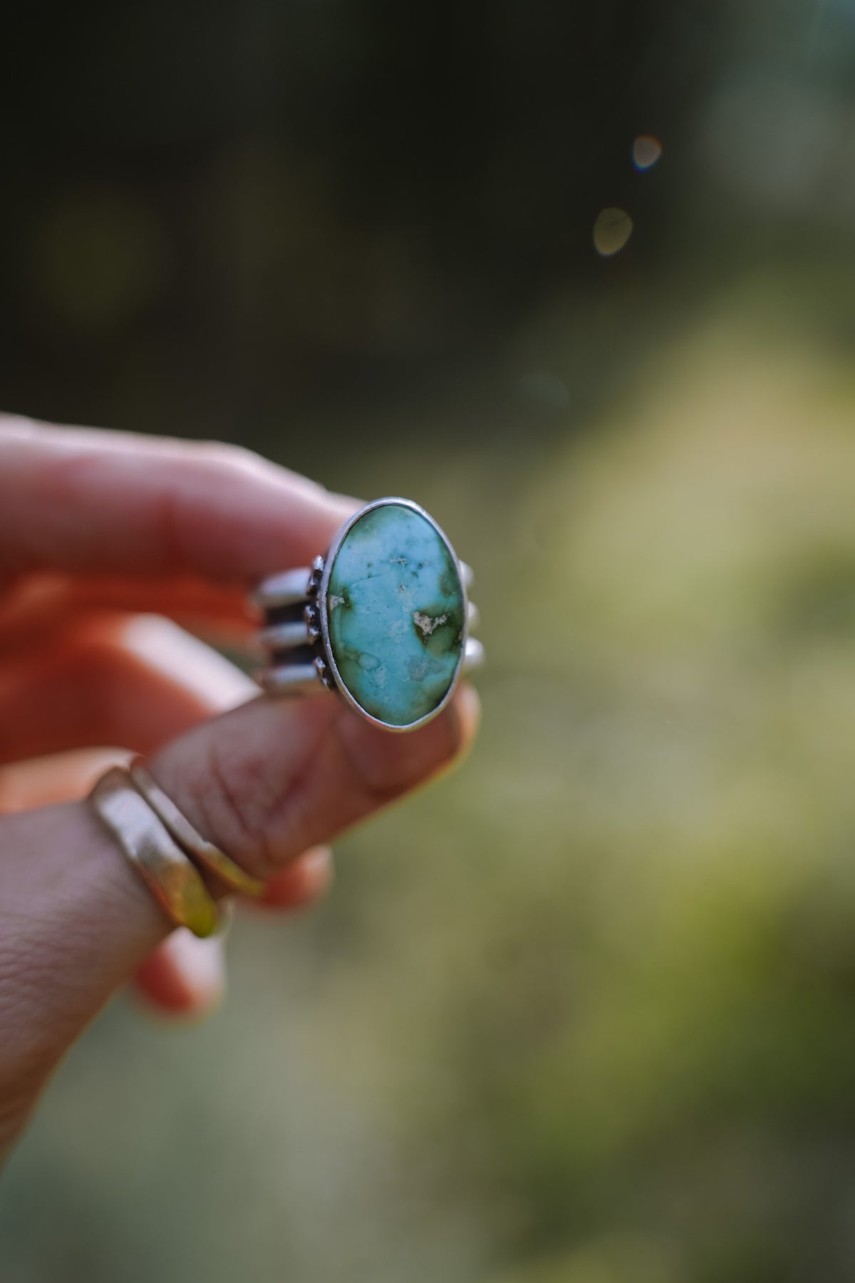 The Cruces ring held up between two fingers, front view, with an oval Sonoran Gold turquoise stone in a smooth silver bezel. The blue-green stone has darker green and gold-toned matrix markings. Small silver crosses or stars decorate the sides of the extra-wide band, and two thin gold bands are stacked on the finger below, against a soft green and golden blurred background.