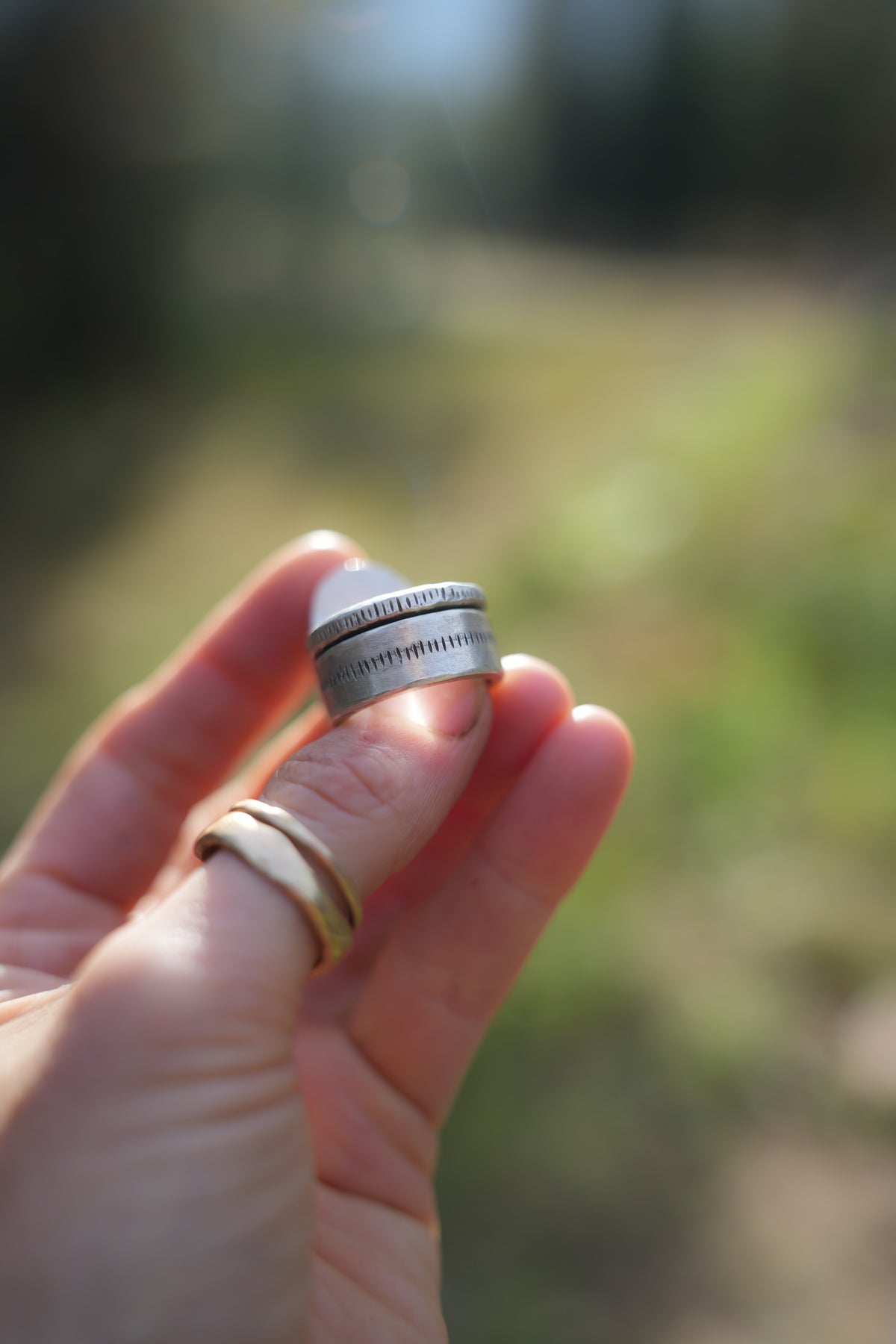 Close-up of the wide band of the Cruces ring, showing two sterling silver bands with fine stamped vertical lines, held between a thumb and finger. The edge of the pale stone is just visible above, with two thin gold bands on the finger below and a blurred green background.