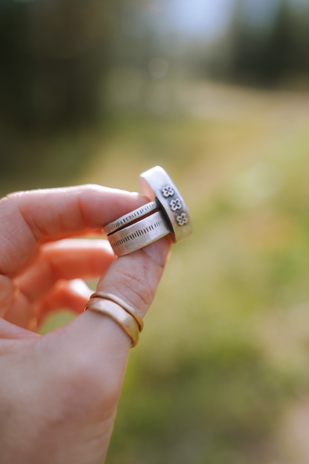 Side view of the Cruces ring held between two fingers, showing a bezel stamped with a row of small four-petal flowers (crosses or stars) above a wide sterling silver band decorated with fine, hand-stamped vertical tick marks. Two thin gold bands sit on the finger below, against a blurred green and golden background.