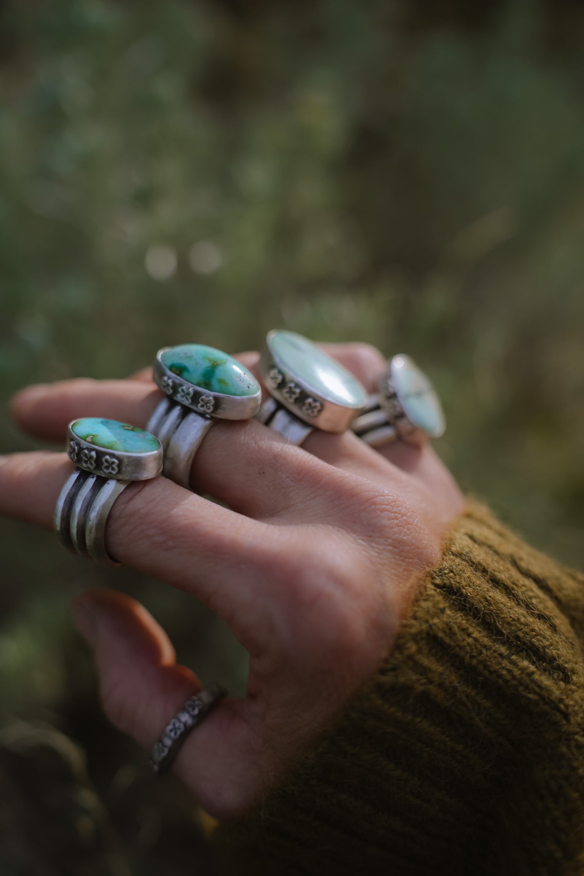 A hand with four sterling silver rings featuring oval turquoise and variscite stones in bezels stamped with small flowers or crosses or stars, set on wide triple-ridge bands, with a matching flower-stamped silver band on the thumb, against a blurred green background and an olive knit sleeve.