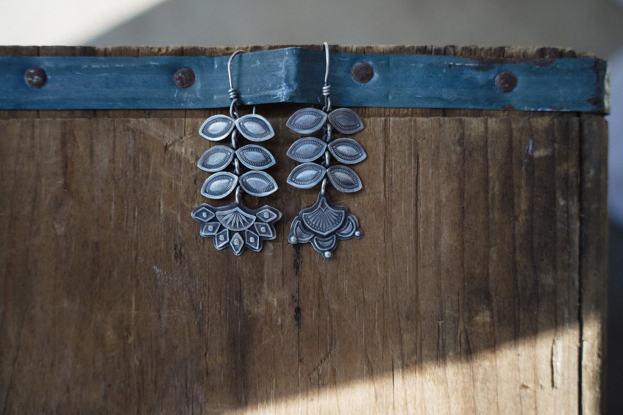 A pair of sterling silver Folk Flower earrings hanging from a worn metal band that is wrapped around a piece of wood. The flower blossoms are at the bottom of the earrings, and six leaf petals are above each blossom. The patterns and markings are hand-stamped.