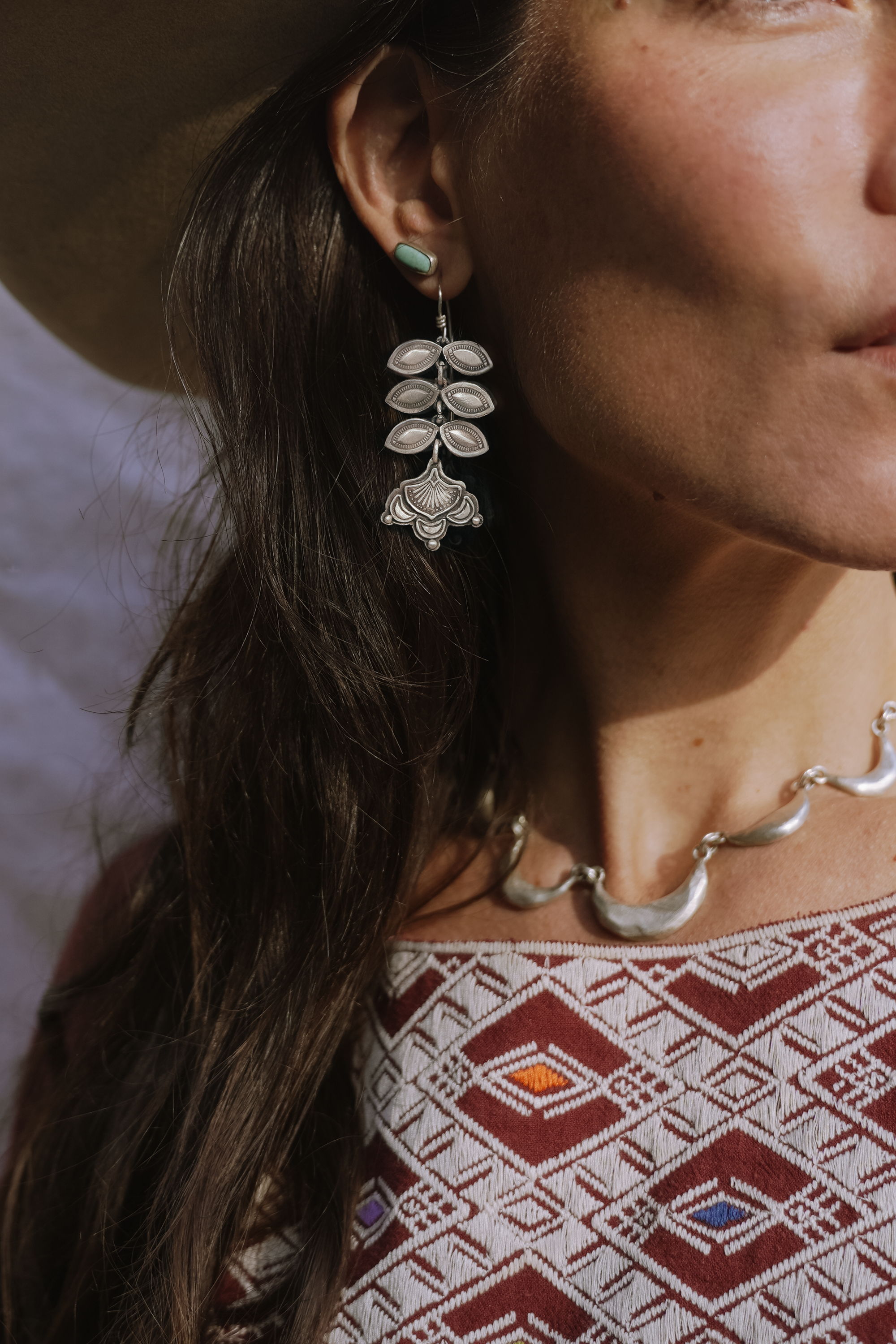 Profile of a woman with long dark brown hair wearing a sterling silver Folk Flower dangling earring and a scalloped sterling silver bracelet. The flower blossom is at the bottom of the earring and six leaf petals are above it.