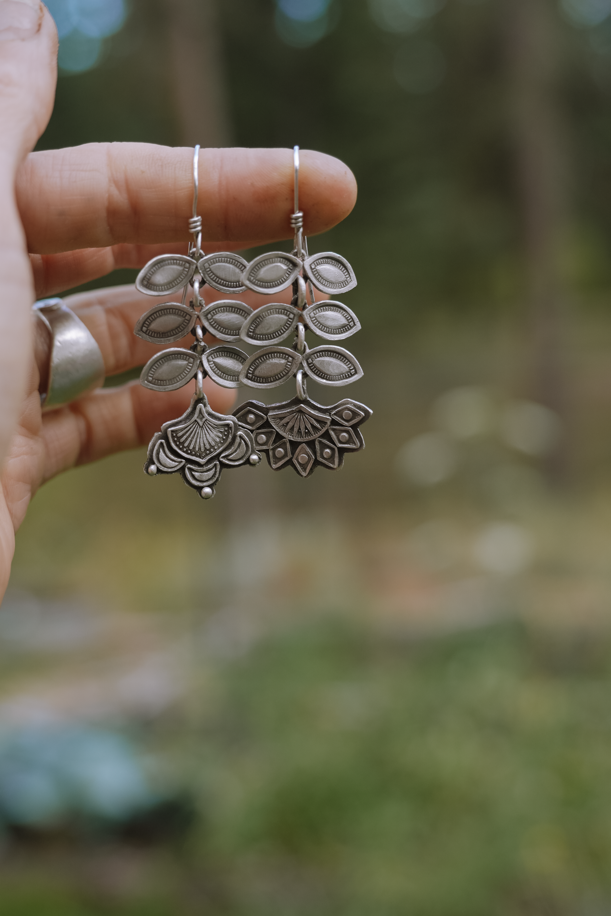 A woman's hand holding a pair of sterling silver Folk Flower earrings. The flower blossoms are at the bottom of the earrings and six leaf petals are above each blossom. The patterns and markings are hand-stamped.