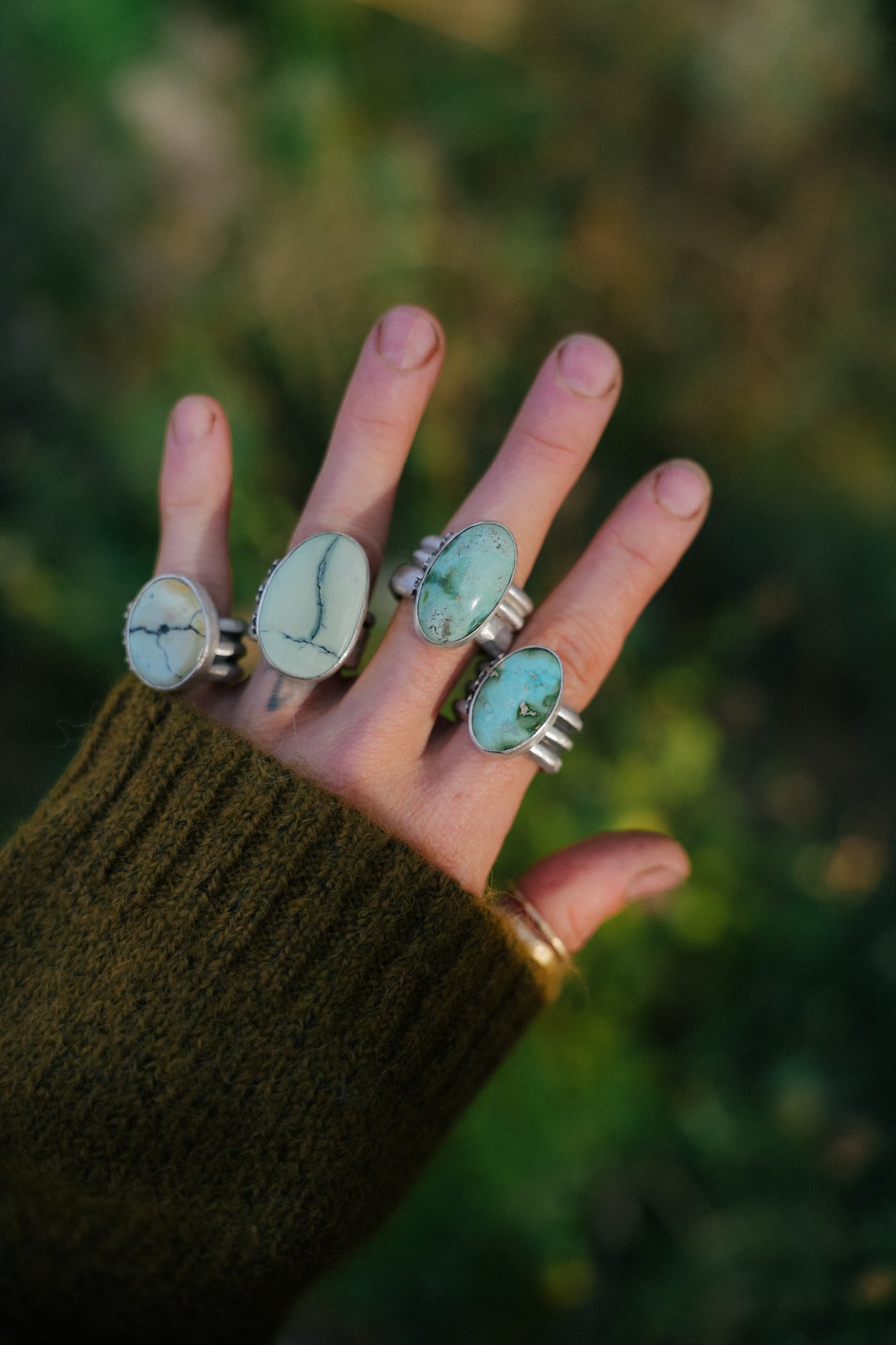 An open hand with four sterling silver rings with large oval stones, two pale green-white variscite with dark veining on the left and two blue-green turquoise on the right, each on a wide triple-ridge band, against a green outdoor background with an olive knit sleeve at the left.