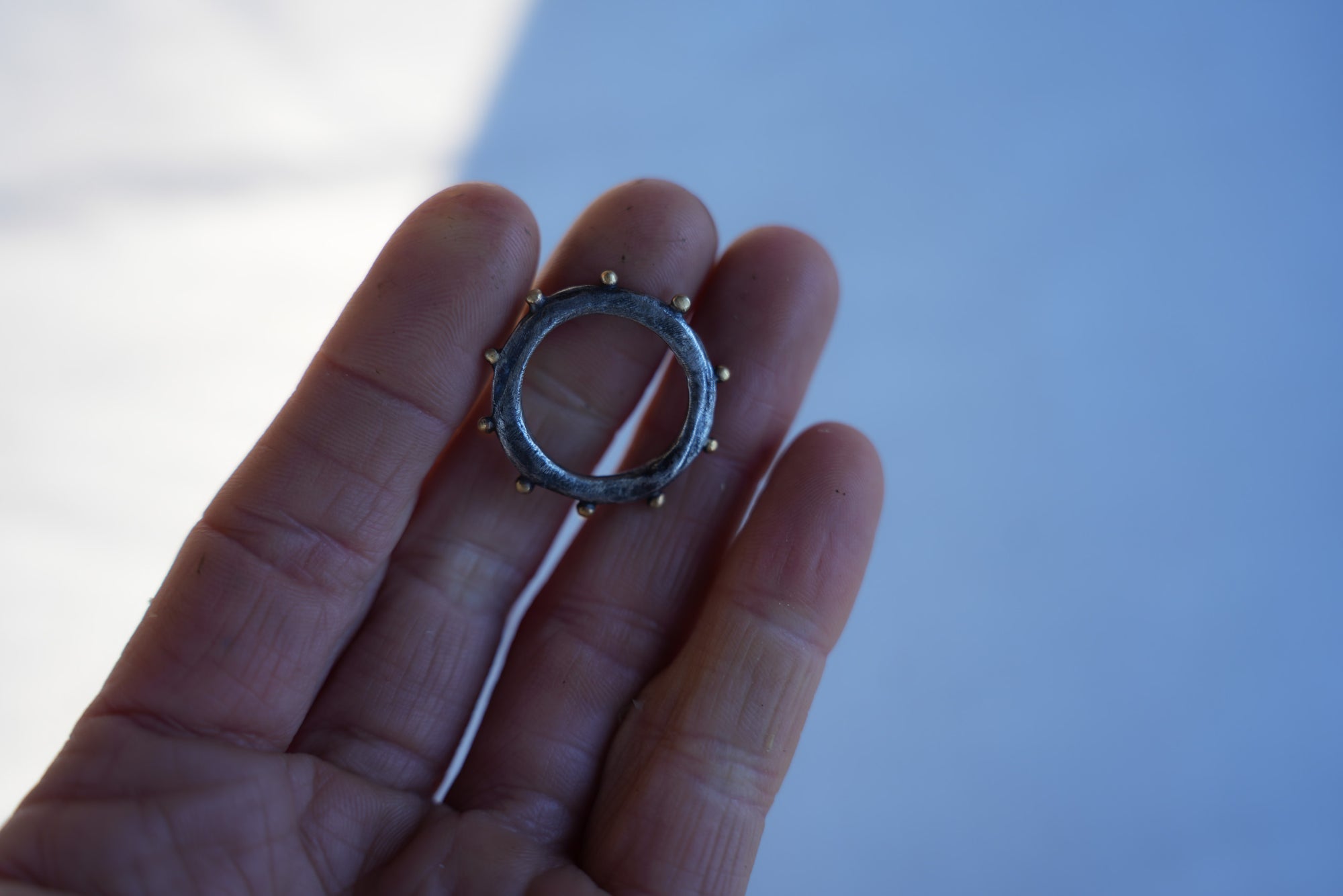 Close-up of a woman's hand. She holding a sterling silver ring with 18K gold beads around the circumference against a blurred background.