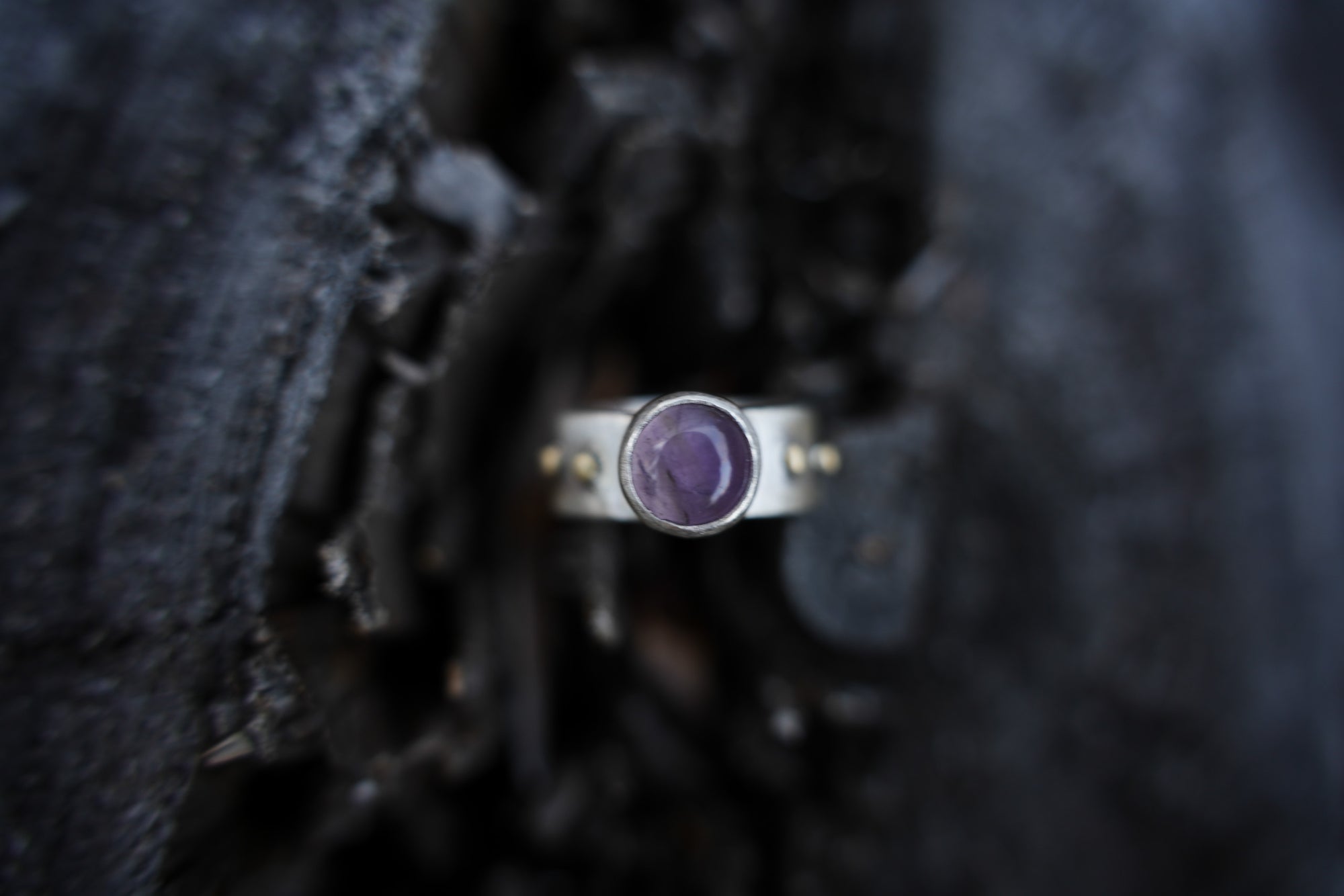 A sterling silver and 18K gold ring with a purple amethyst gemstone against a dark, blurred background.