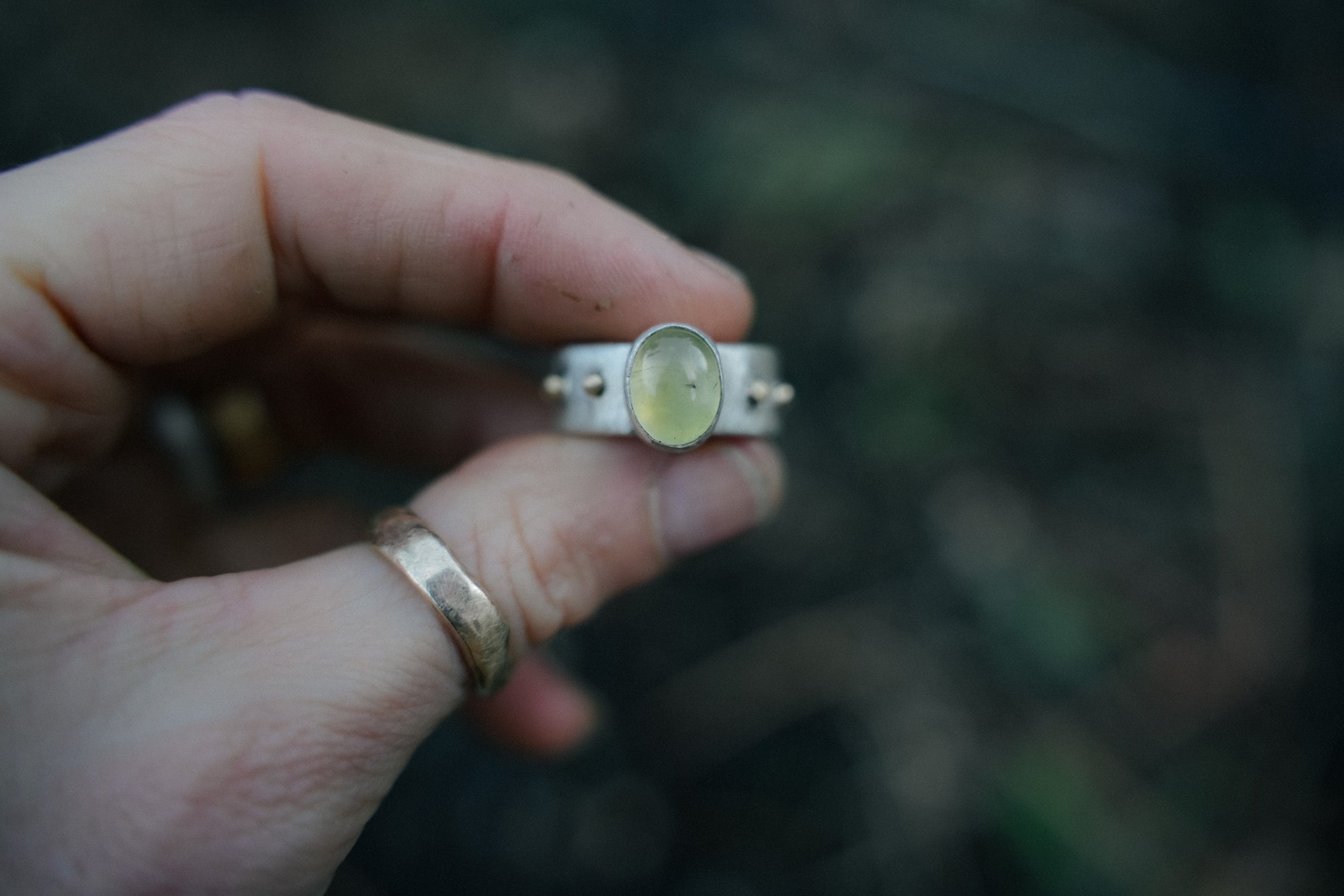 A woman’s hand holding a sterling silver and 18K gold ring with a light green citrine gemstone against a blurred background.