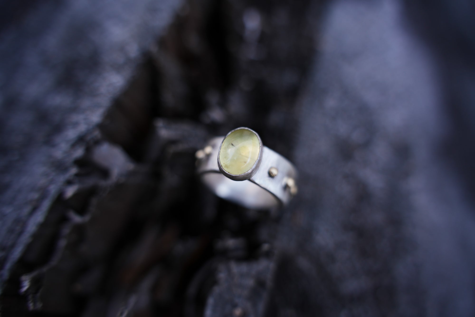 A sterling silver and 18K gold ring with a light green citrine gemstone against a dark, blurred background.