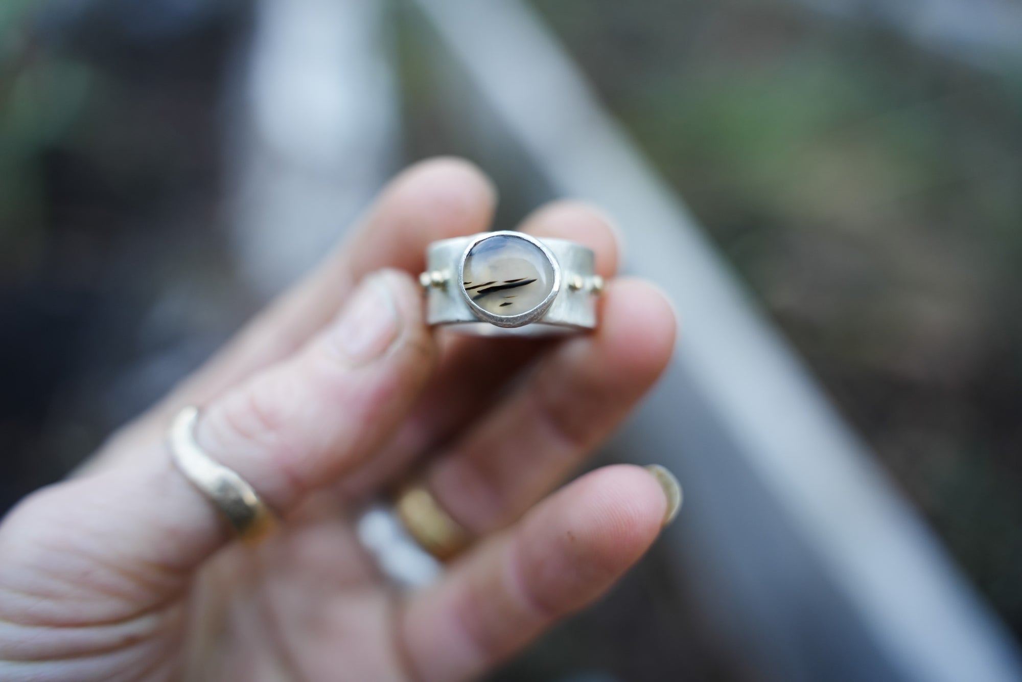 Close-up of a woman's hand. She holding a sterling silver and 18K gold ring with a Montana agate against a blurred background.