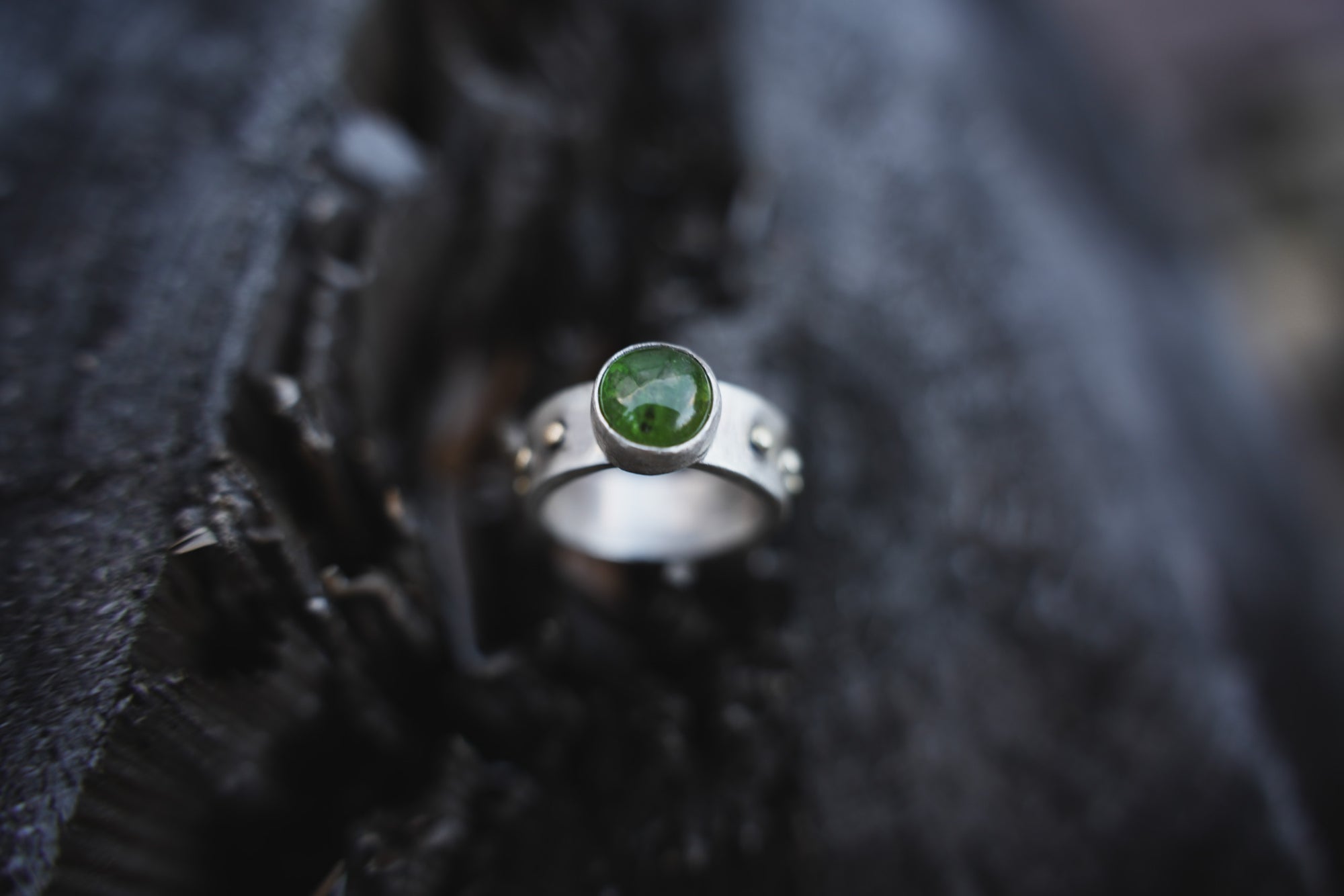A sterling silver and 18K gold ring with a dark green vesuvianite gemstone against a dark, blurred background.
