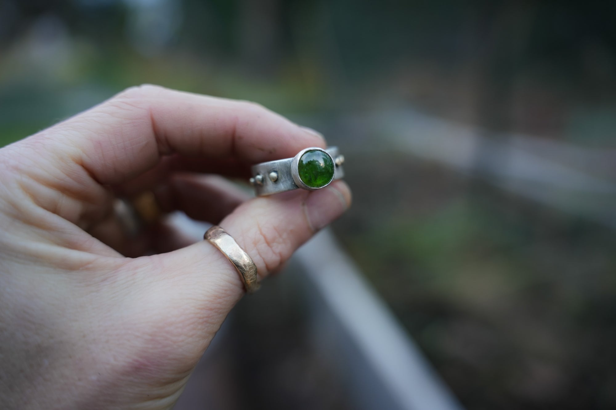Close-up of a woman's hand. She holding a sterling silver and 18K gold ring with a dark green vesuvianite gemstone against a blurred background.