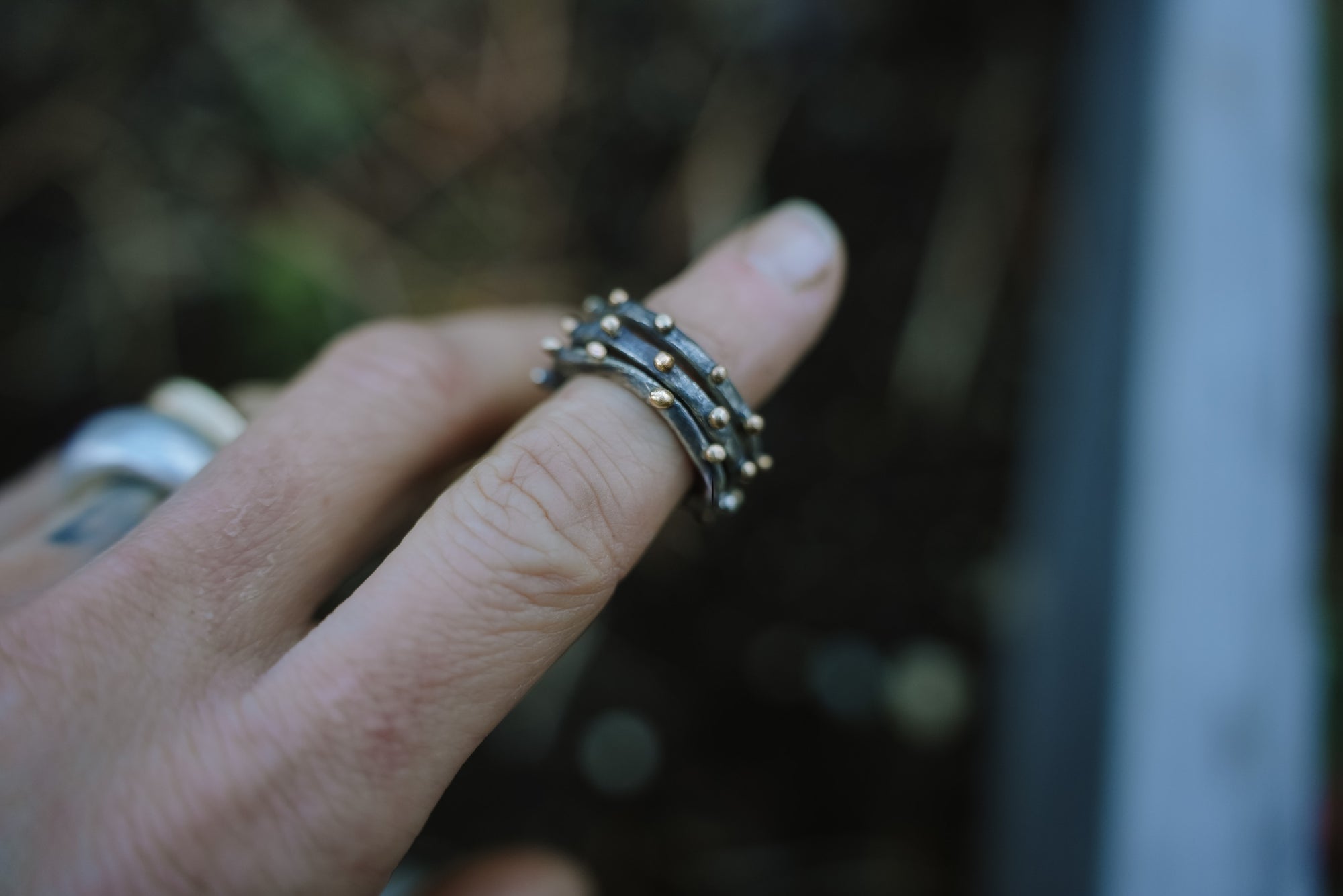 Close-up of a woman's hand. She is wearing a stack of sterling silver and 18K gold rings on her forefinger. The silver has been oxidized, so it is black. The background is blurred.