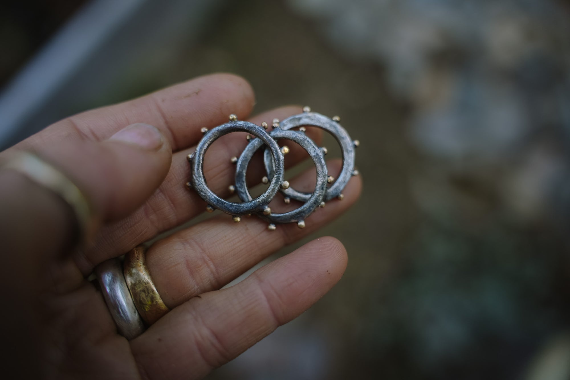 A woman's hand holding three oxidized sterling silver rings with 18K gold beads against a blurred natural background