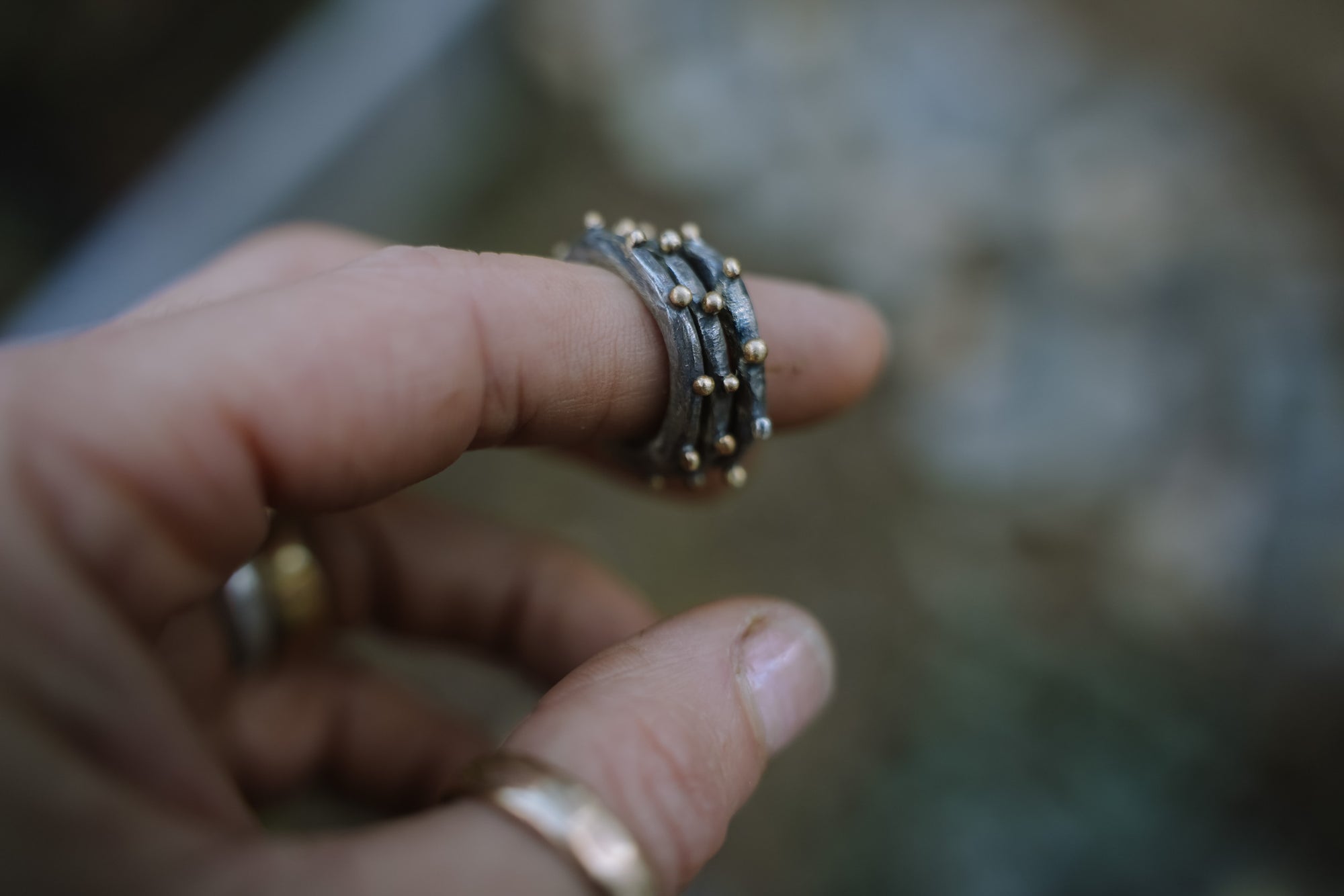 A woman's hand photographed a blurred background. She is wearing three oxidized sterling silver rings with 18K gold beads on her forefinger. 