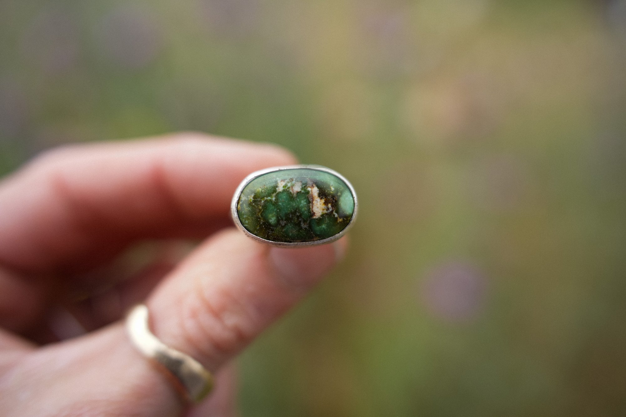 A woman's hand holding a sterling silver ring with an oval green turquoise gemstone which has dark matrix and small gold inclusions, set against a blurred outdoor background.