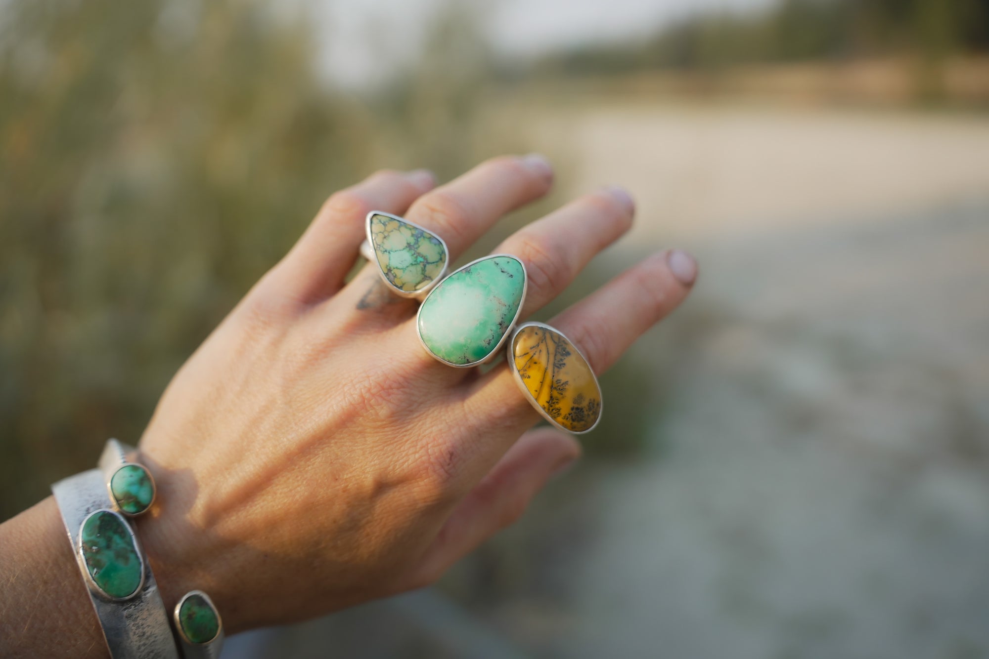 A woman's hand wearing multiple sterling silver rings with green and orange gemstones against a blurred natural background